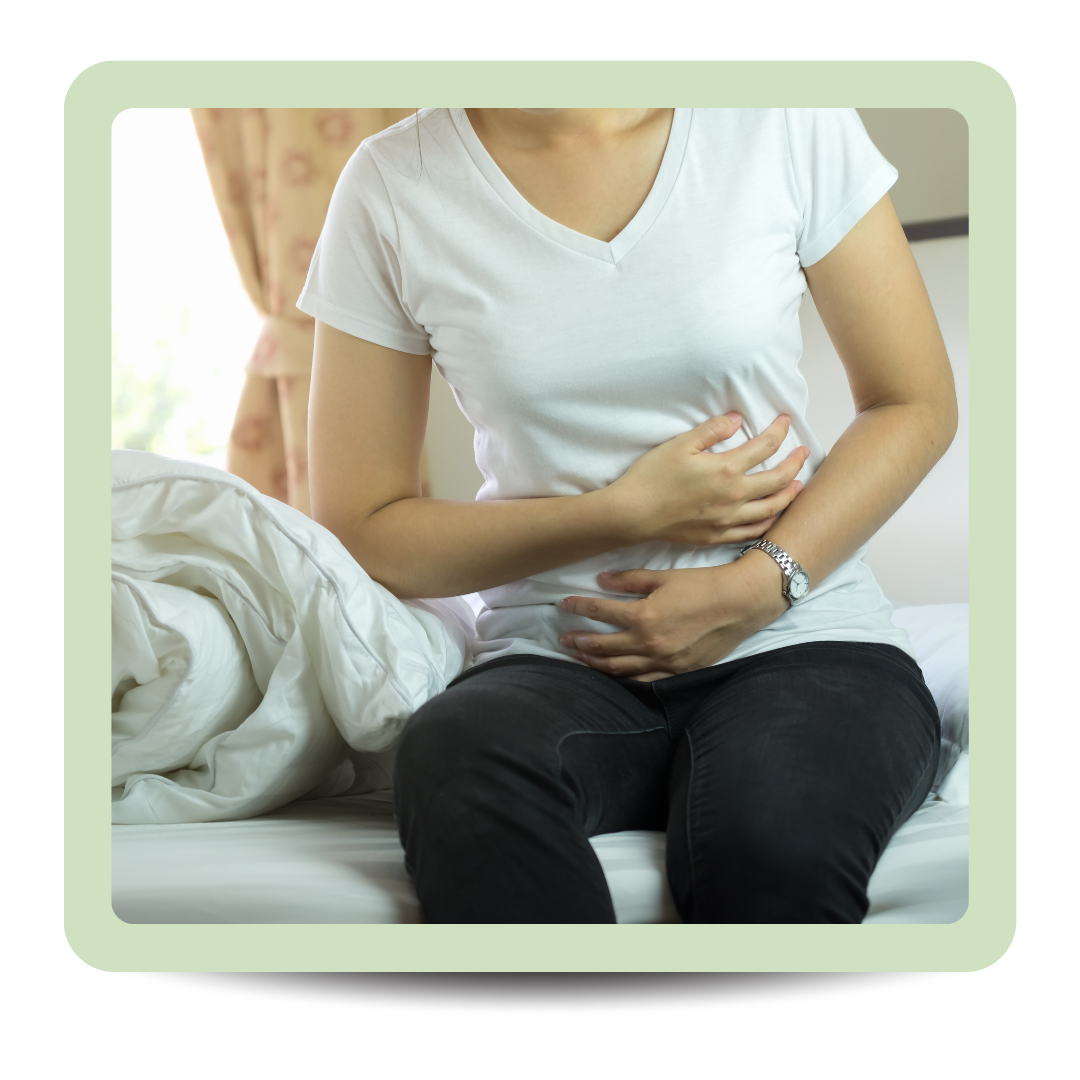 Woman wearing a white t-shirt and black pants sitting on bed, clutching her stomach with one hand, indicating stomach pain or discomfort.