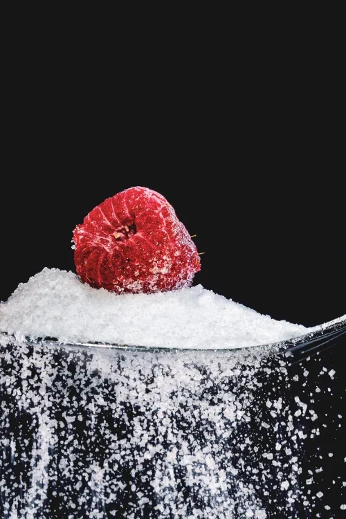 A raspberry on a mound of sugar in a clear glass container against a black background.