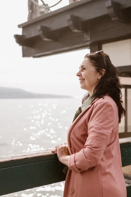 A smiling woman standing on a boat, looking out at the water on a bright day.