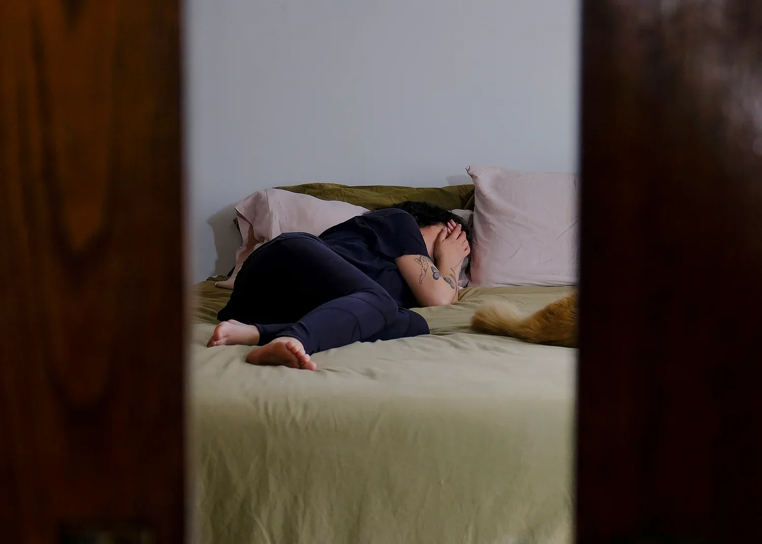 Person lying on bed, curled up and covering face with hands, seen through door frame