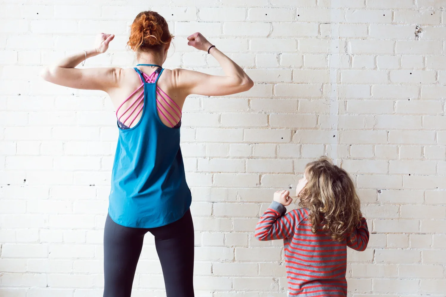 A woman flexing her arms and showing her muscles while a young girl looks up at her. They are standing against a white brick wall.
