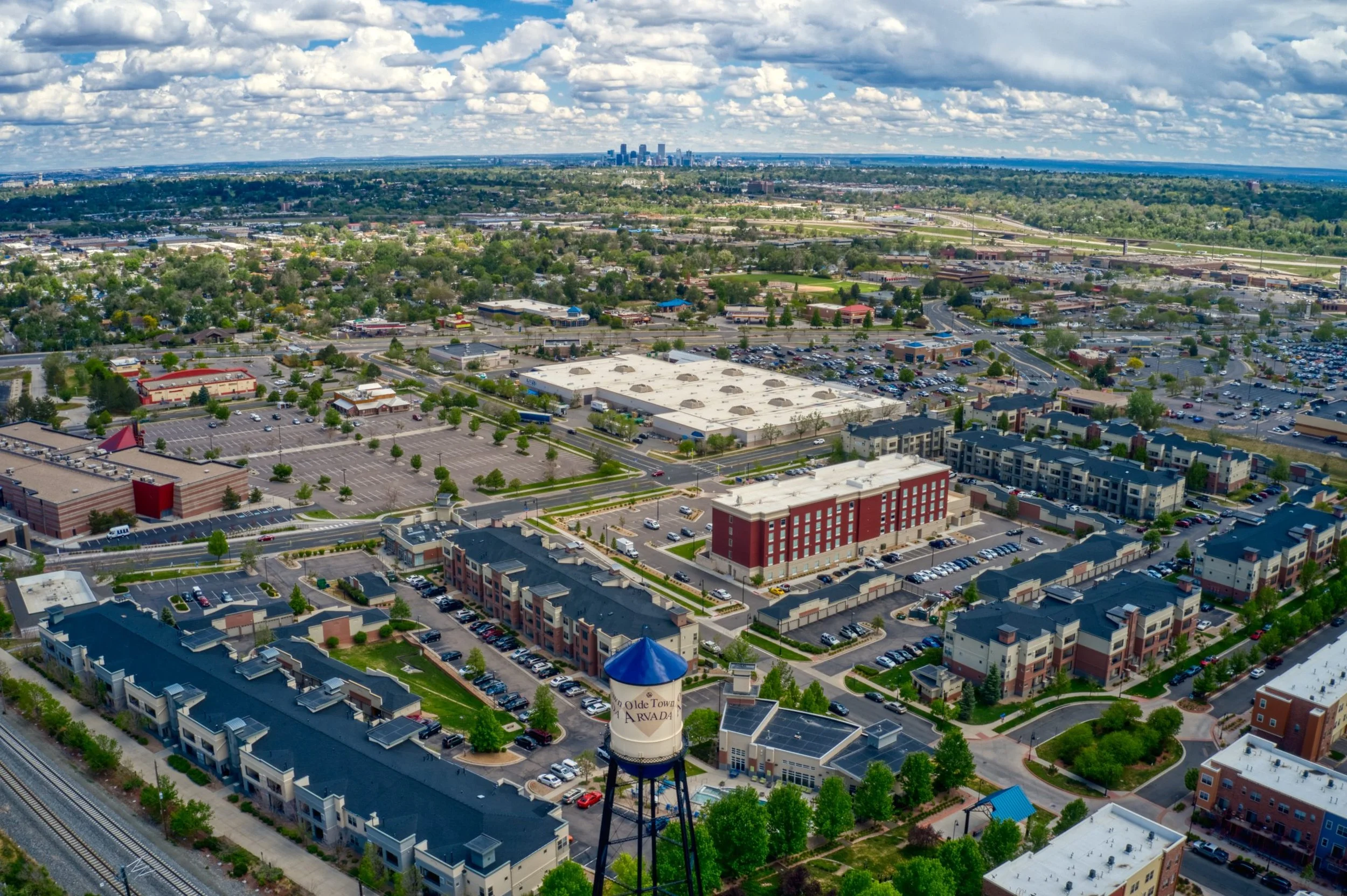 Aerial view of a cityscape with a water tower, commercial and residential buildings, parking lots, and lush greenery, with a distant skyline of tall buildings underneath a partly cloudy sky.