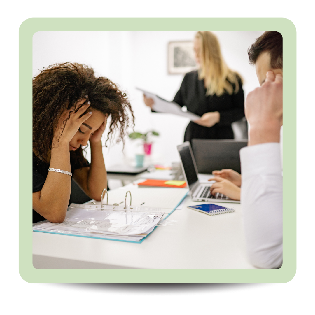 Concerned woman with curly hair sitting at a desk with binders and a laptop, looking stressed in an office with two colleagues in the background.