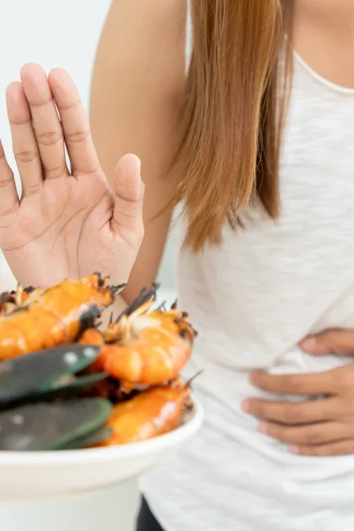 A woman holding up her hand in a 'stop' gesture near a plate of cooked shrimp, with her other hand on her hip.