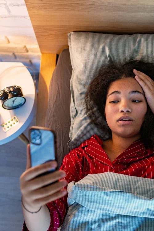 A young woman lying in bed and looking at her phone with a concerned expression, with an alarm clock and medicine on a bedside table.