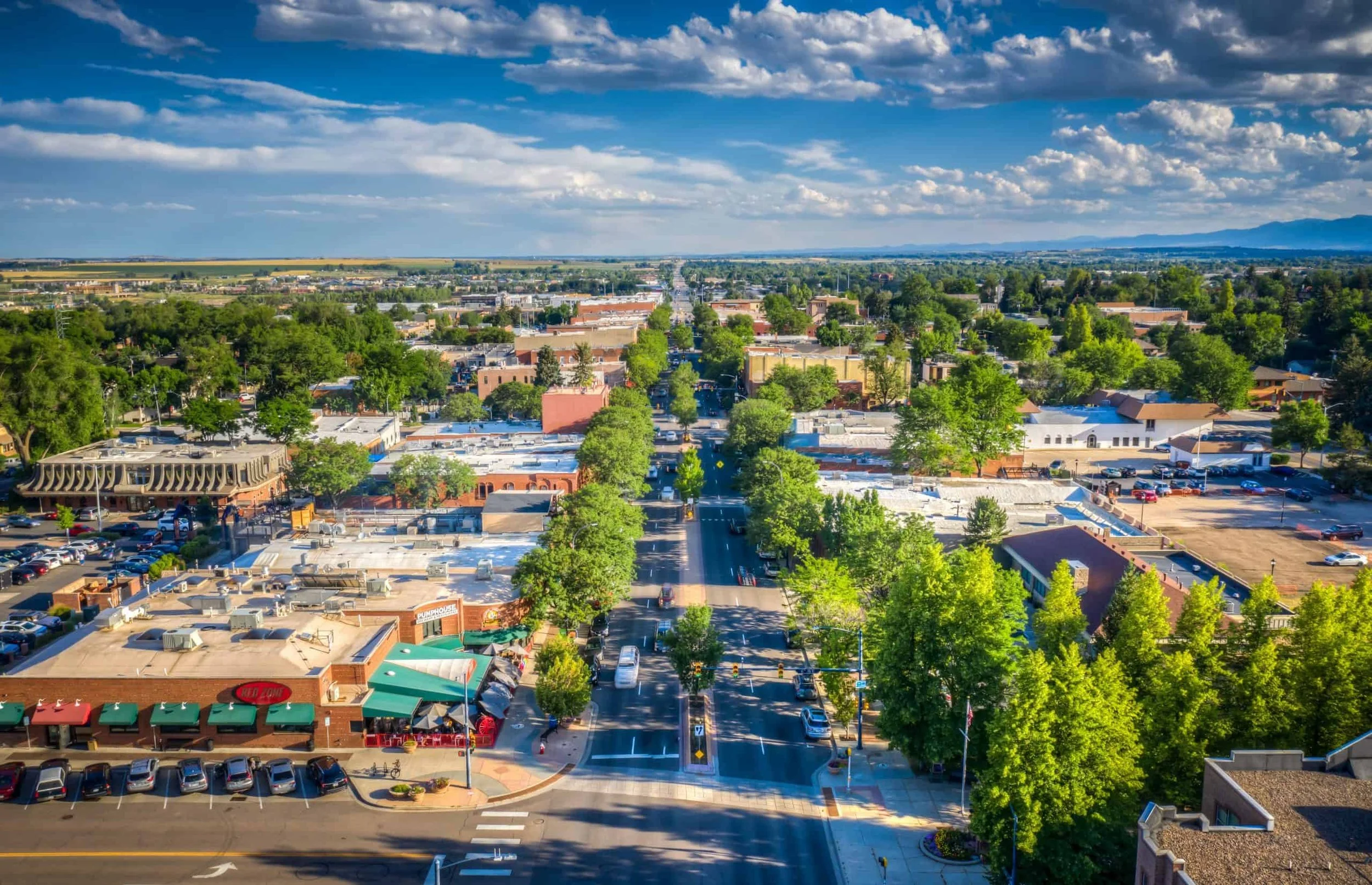 An aerial view of a small town under a partly cloudy sky, with streets, trees, buildings, and parked cars visible.