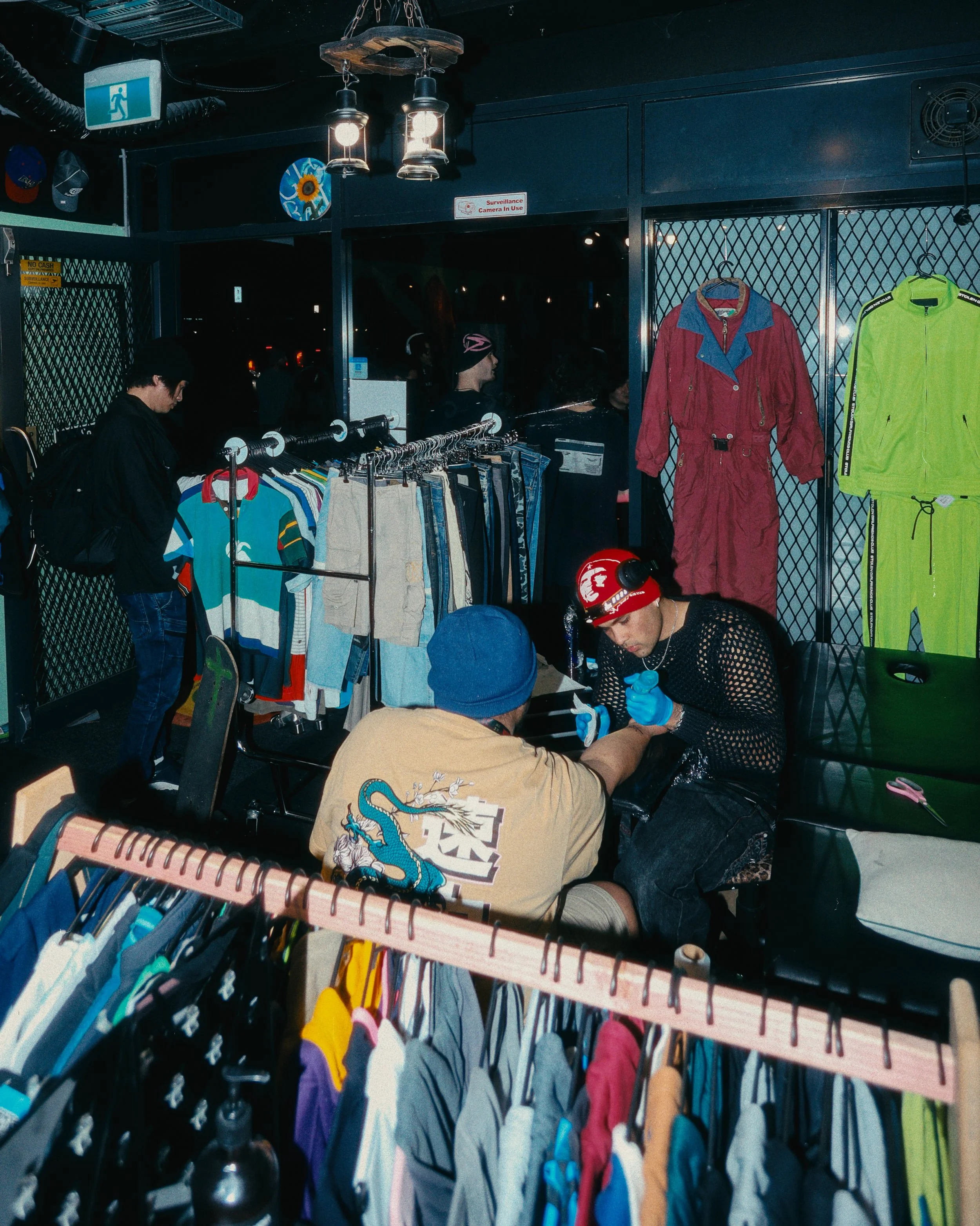People shopping for clothes inside a thrift store; racks of colorful clothing, jackets, and shirts; individuals browsing and examining items; dark interior with hanging lights. A person is getting tattooed in this space.