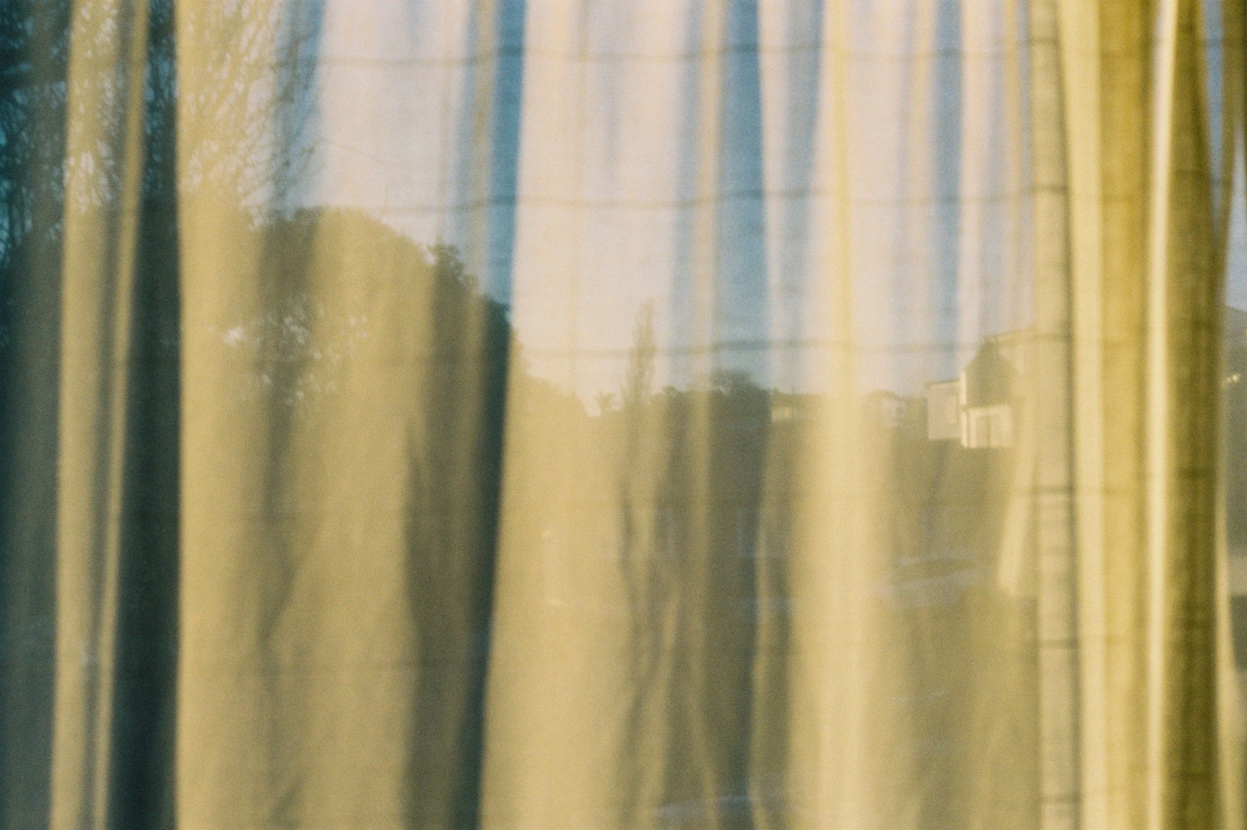 The image shows a view through a window with semi-transparent yellow curtains, with some trees and houses visible in the background.