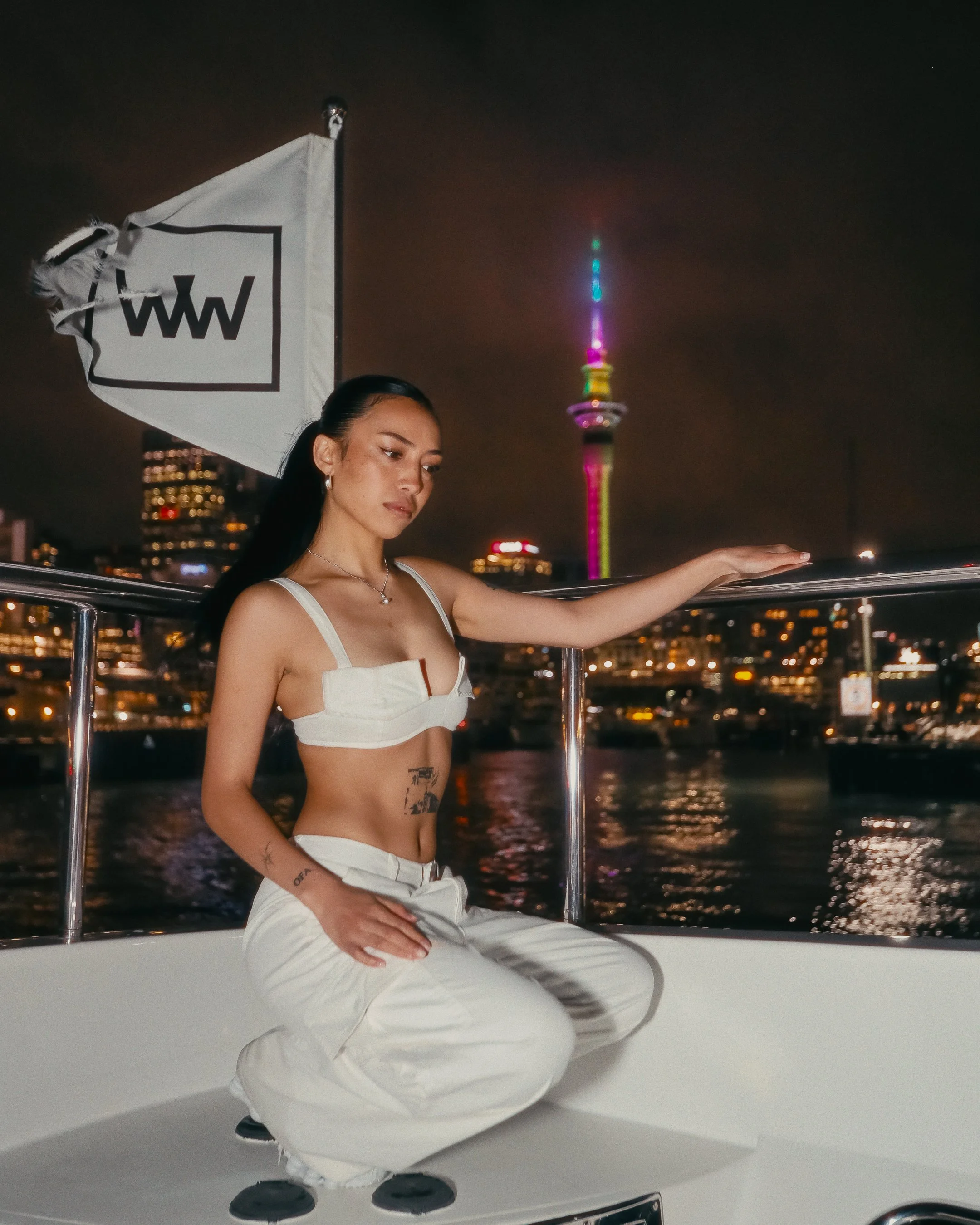 A woman with long dark hair stands on a boat at night, with Auckland city lights and the Sky Tower in the background. She is dressed in white pants and a white cropped top, leaning on the boat's railing.