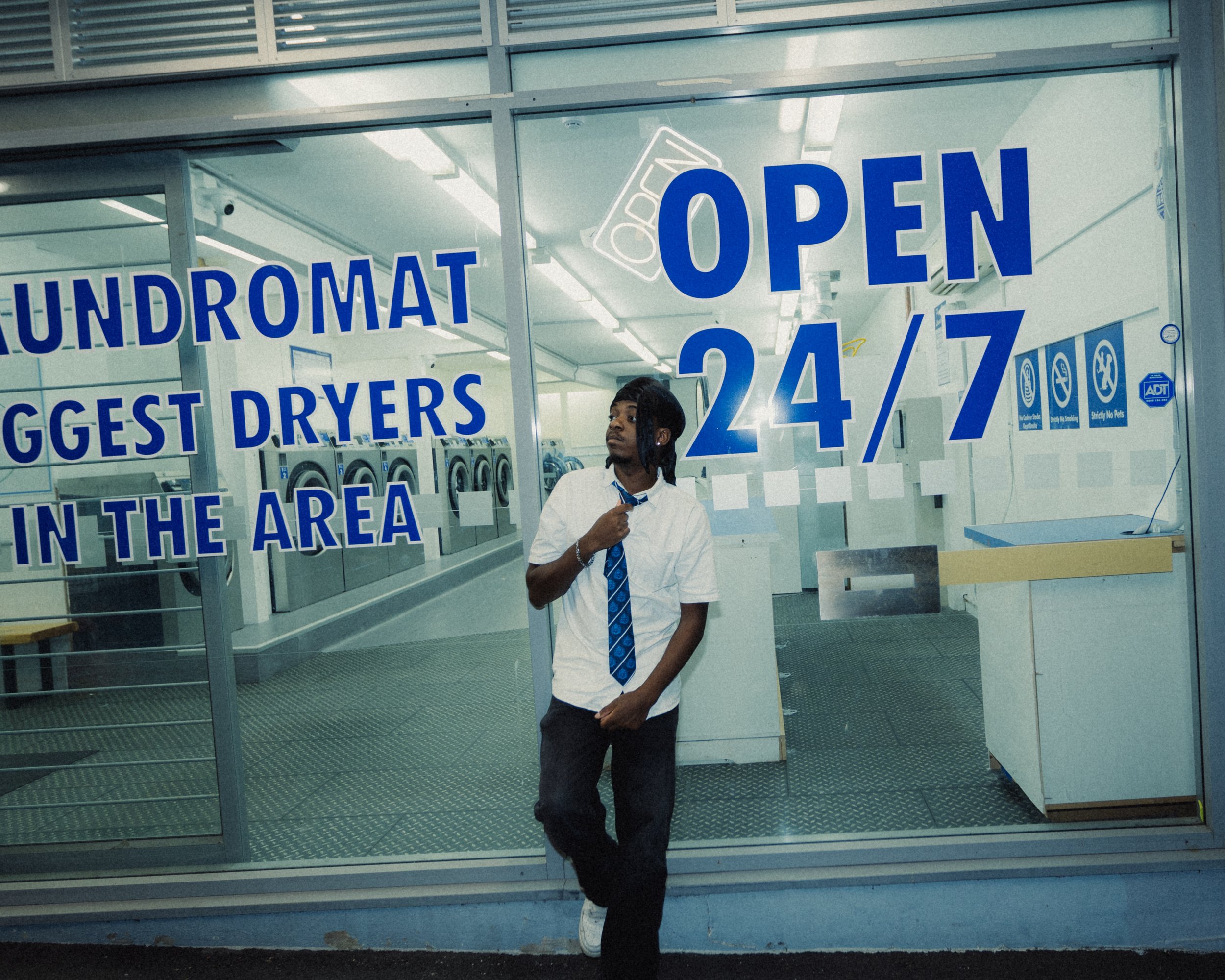 A person wearing a white shirt, blue tie, and durag standing in front of a laundromat window that has a sign that reads 'LAUNDROMAT OPEN 24/7' 