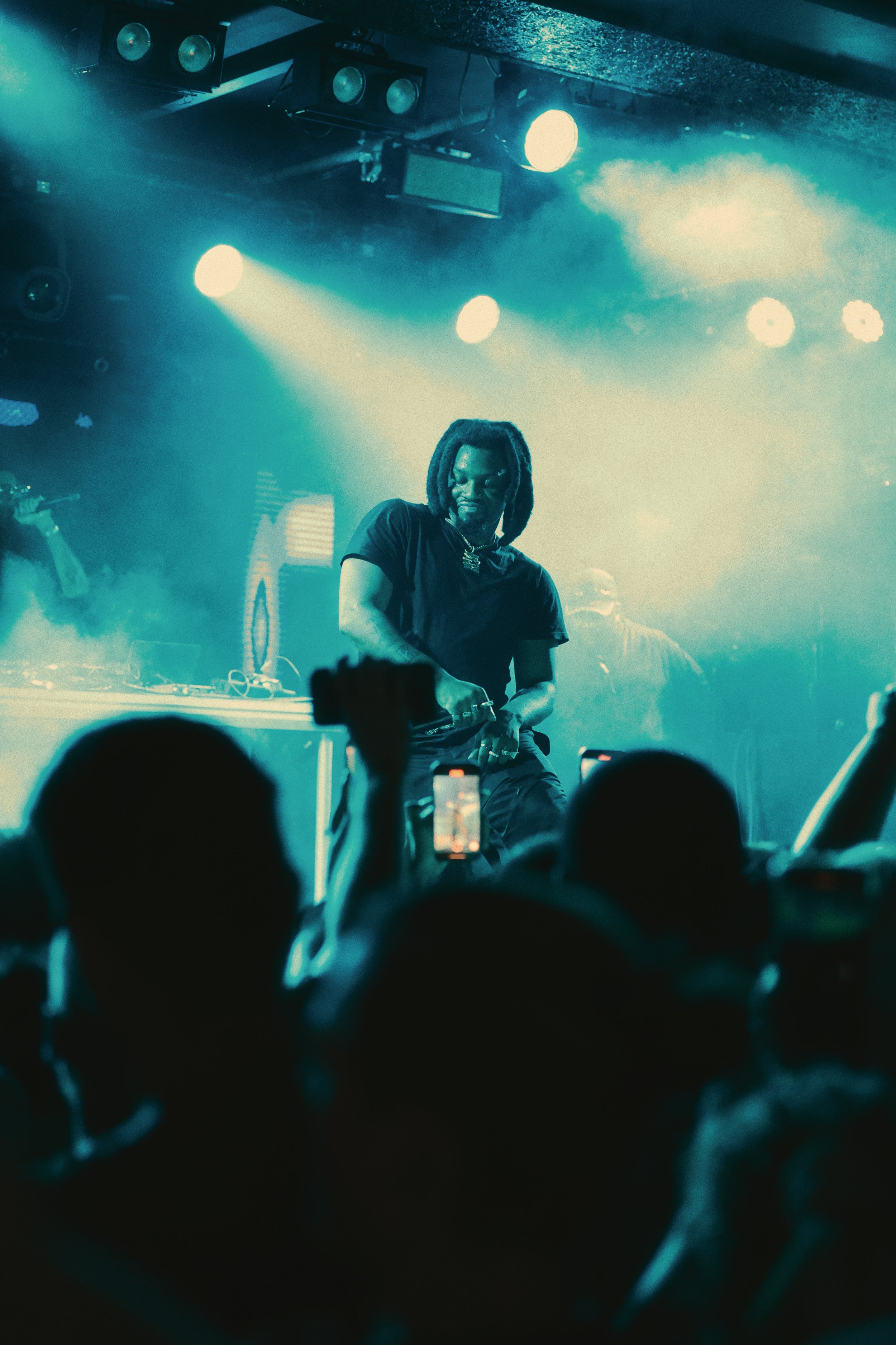 Denzel Curry performing on stage, with blue lights beaming as the audience takes photos.