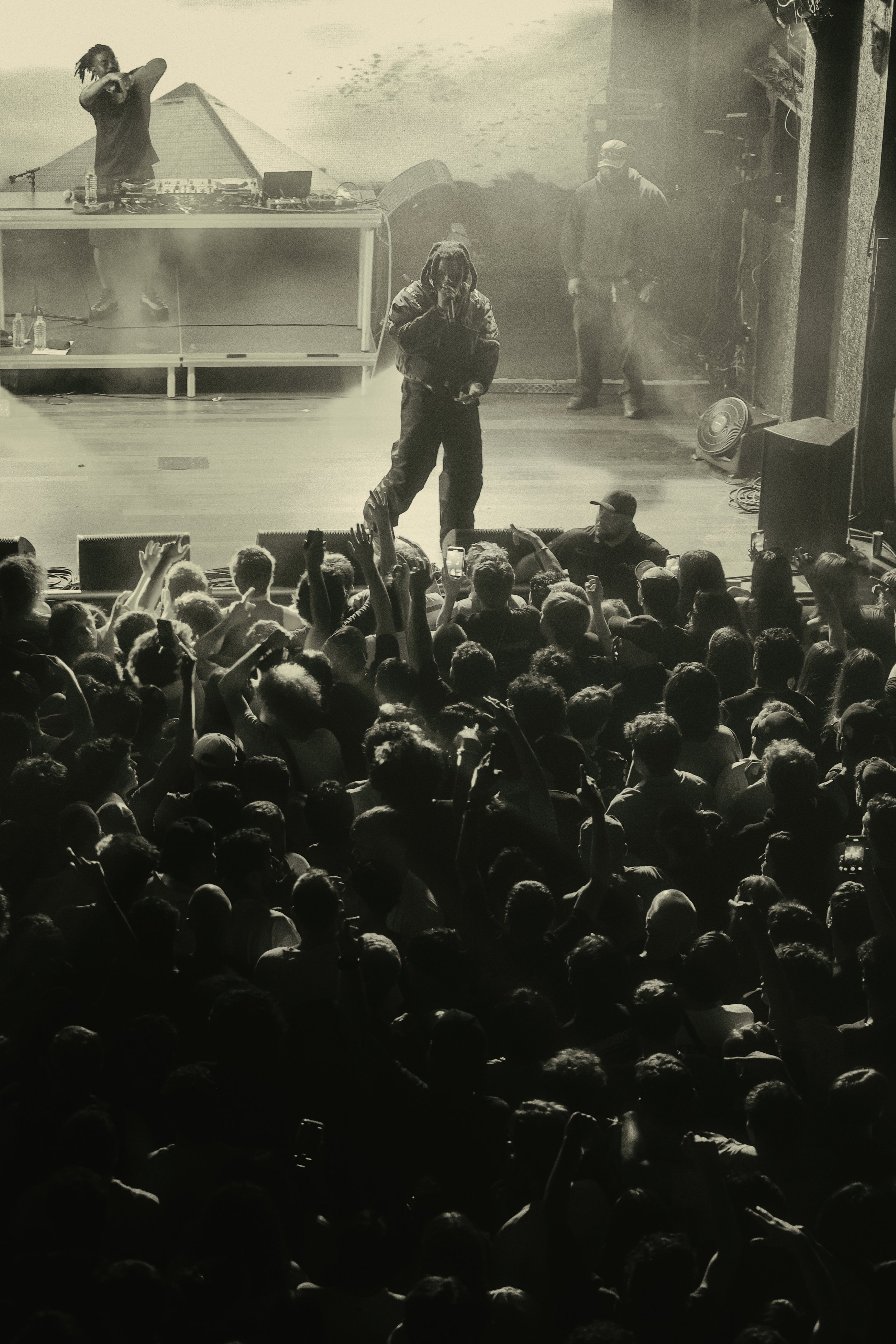 A black and white photo of a Denzel Curry concert with a DJ in the background, and a large audience with many raised hands.