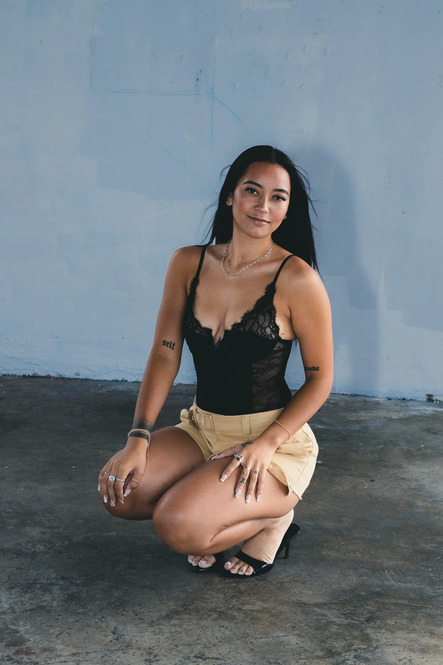 A portrait of a young woman with long black hair, wearing a black lace top and beige shorts, is crouched on the ground against a gray concrete wall with a slight smile, accessorized with rings, necklaces, and bracelets.