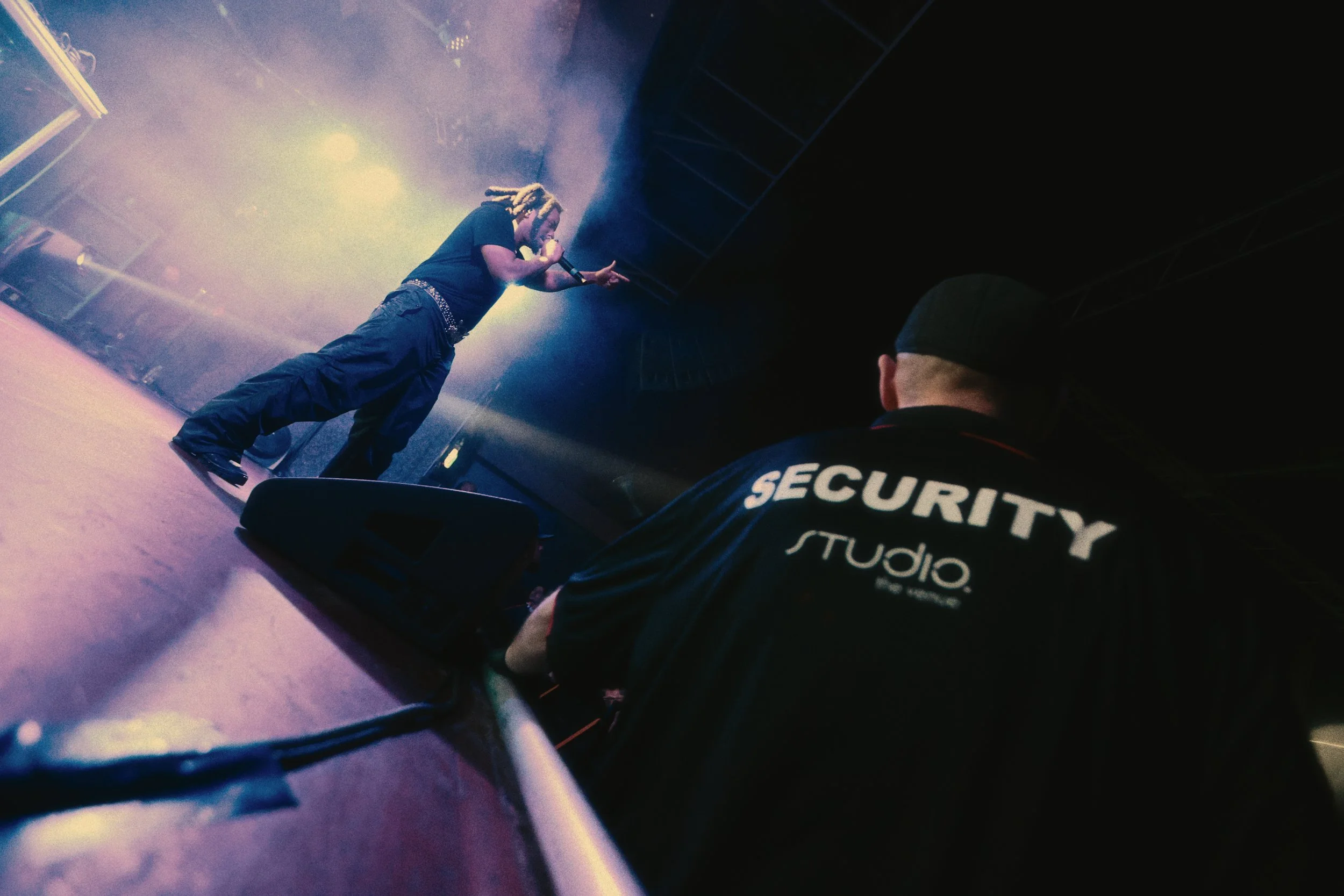 Denzel Curry singing into a microphone on stage, with colorful stage lights and smoke in the background. A security guard is in the foreground.