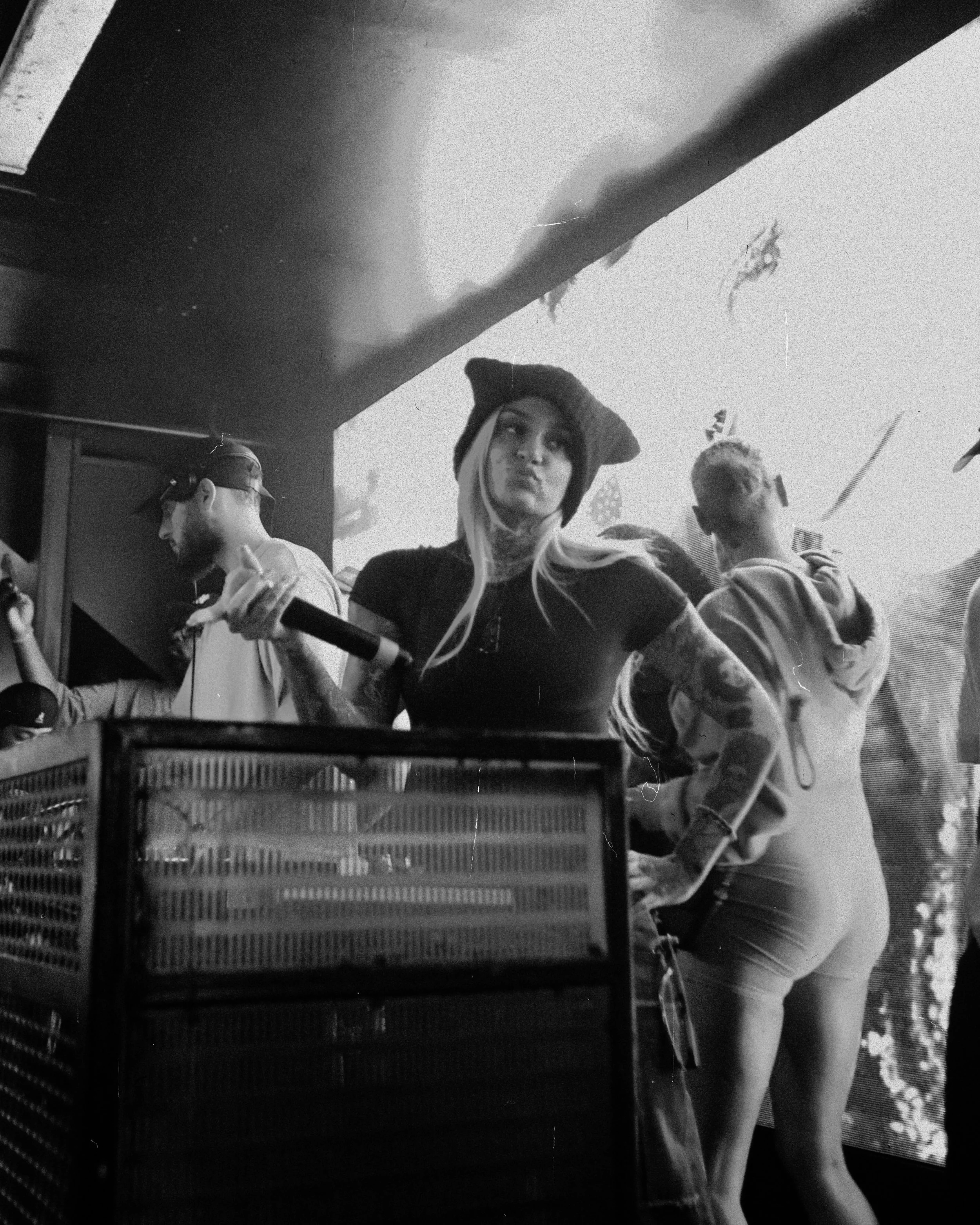 A black and white photo of Kehlani wearing a beanie, standing behind a DJ booth, with multiple people in her entourage dancing around her. The woman's pose and expression suggest she is performing.
