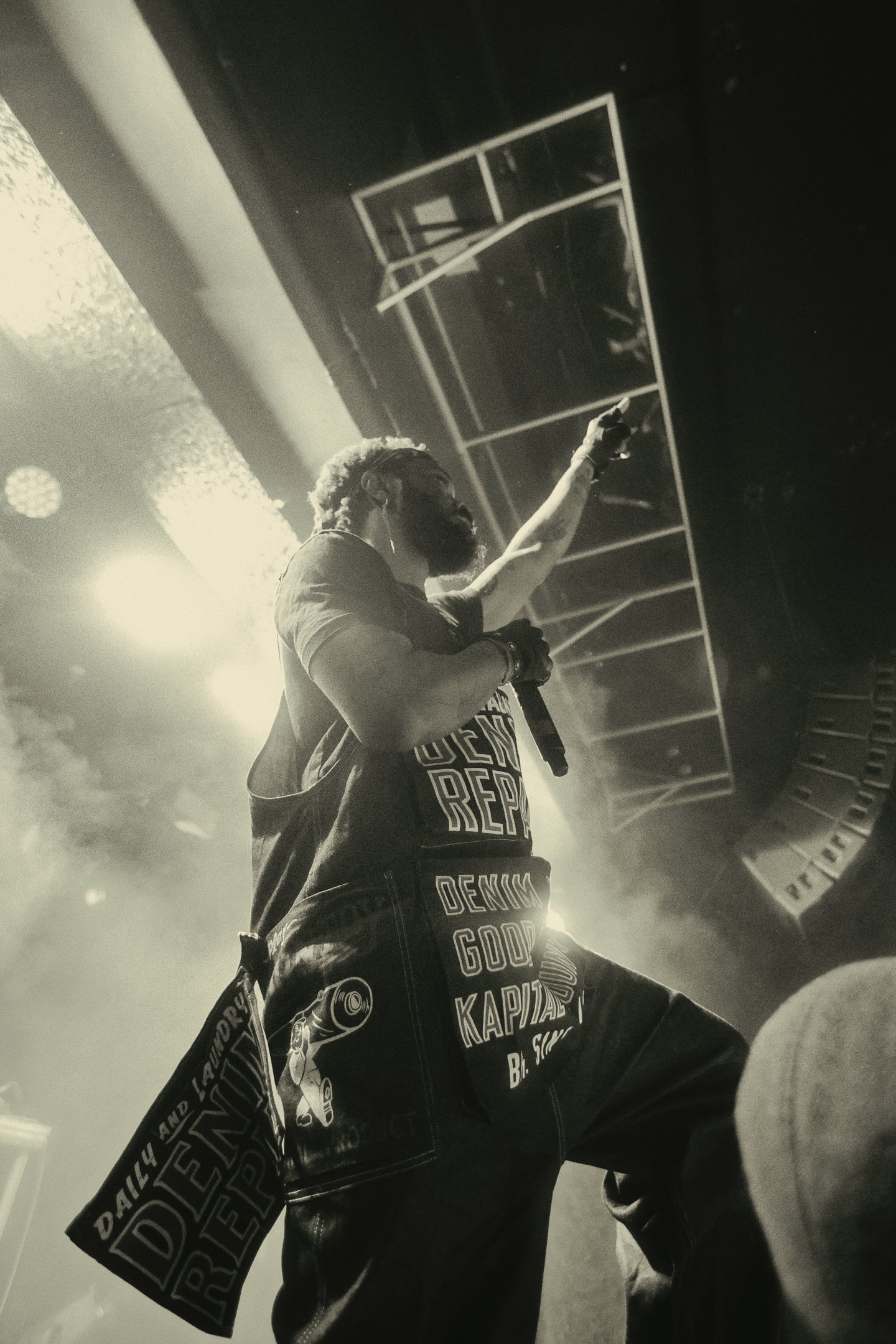 JPEGMAFIA on stage wearing a black t-shirt and jeans, left fist in the air. The stage has lighting effects and audio equipment, and the photo is taken from a low angle.