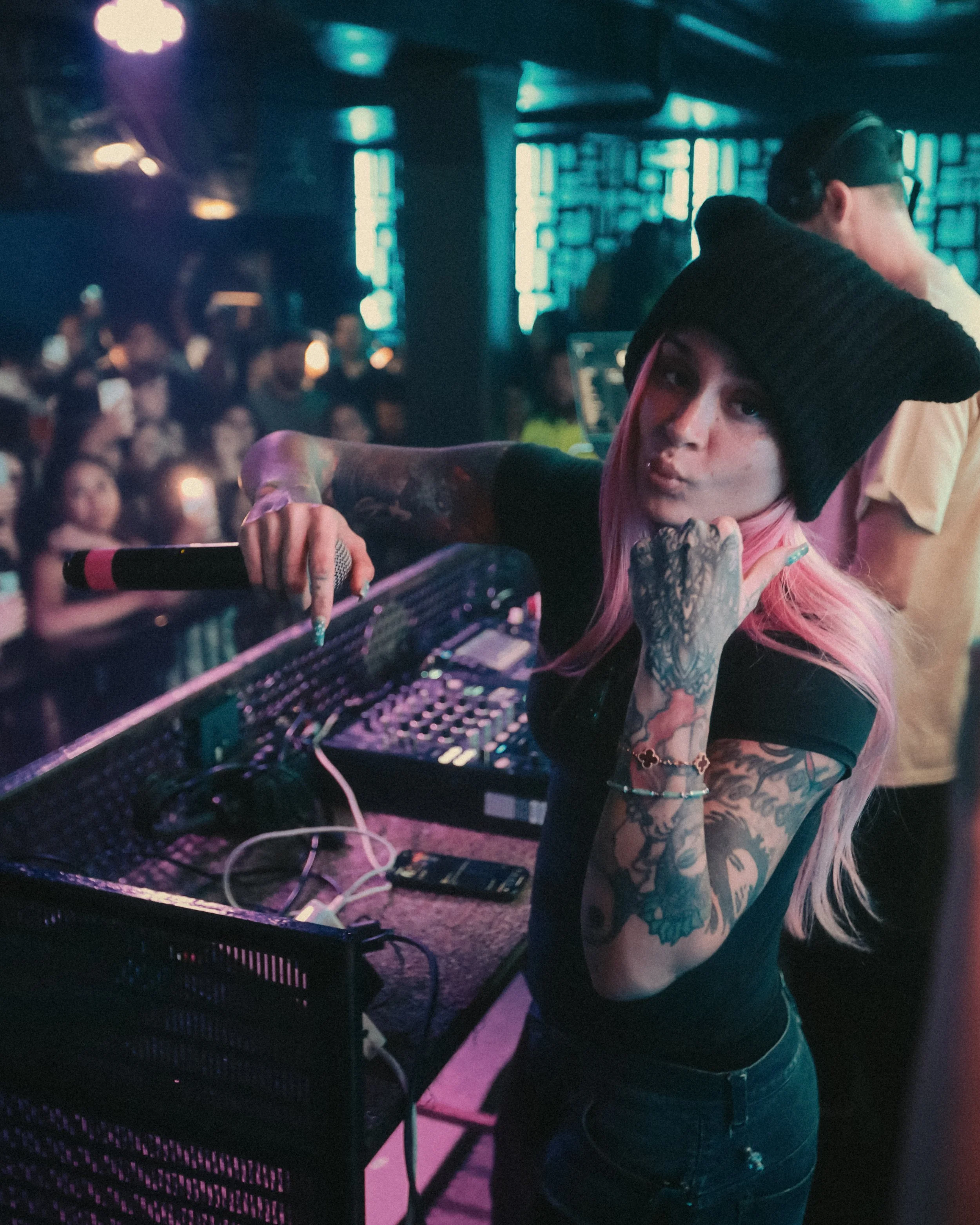 Kehlani with pink hair and tattoos is at a club performing, being the DJ booth, wearing a black knit hat and black shirt, looking back at the cameras people dance in the background.