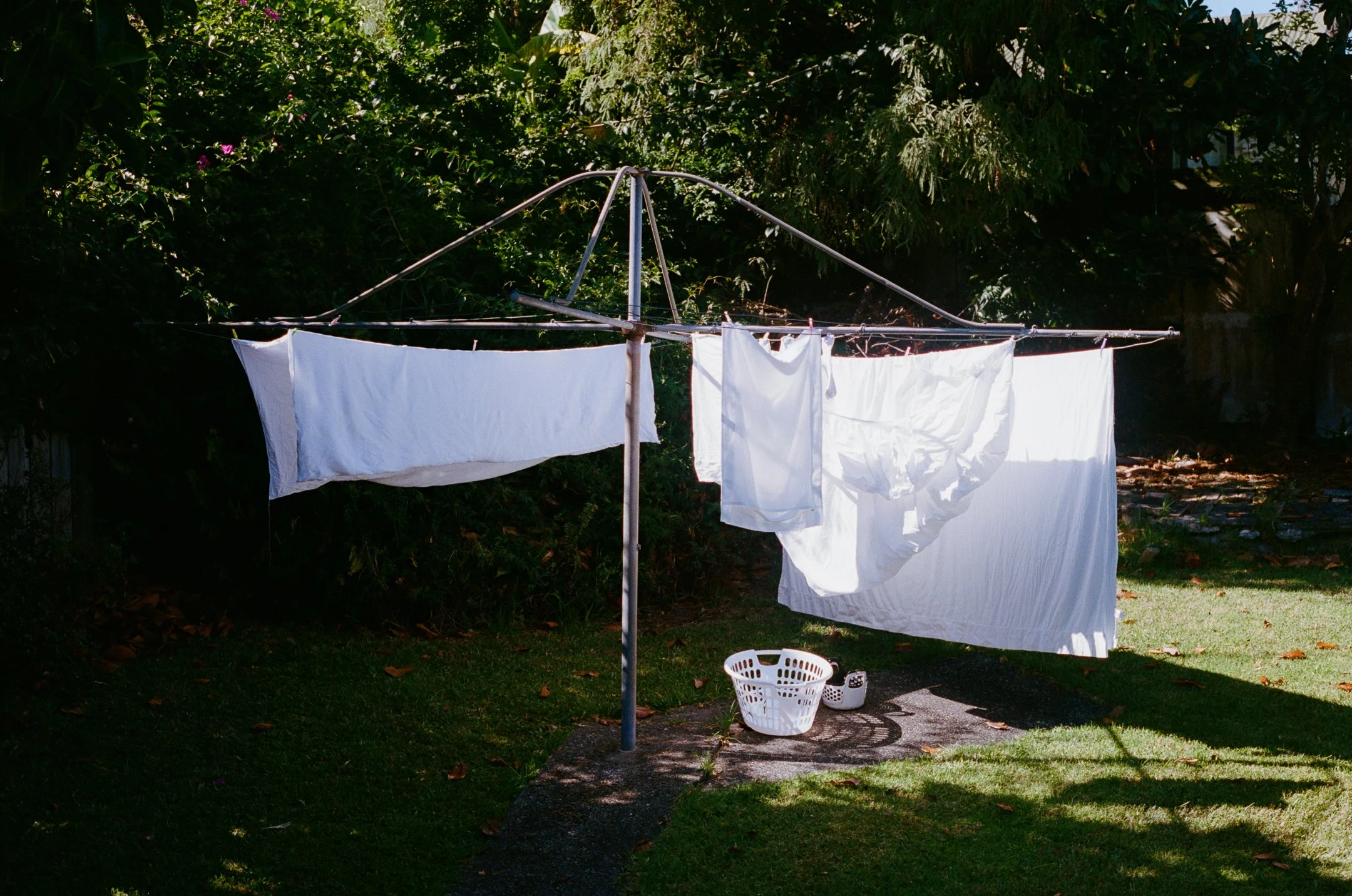 Clothes hanging on a rotary clothesline in a backyard with green bushes and trees, with laundry baskets on the ground.