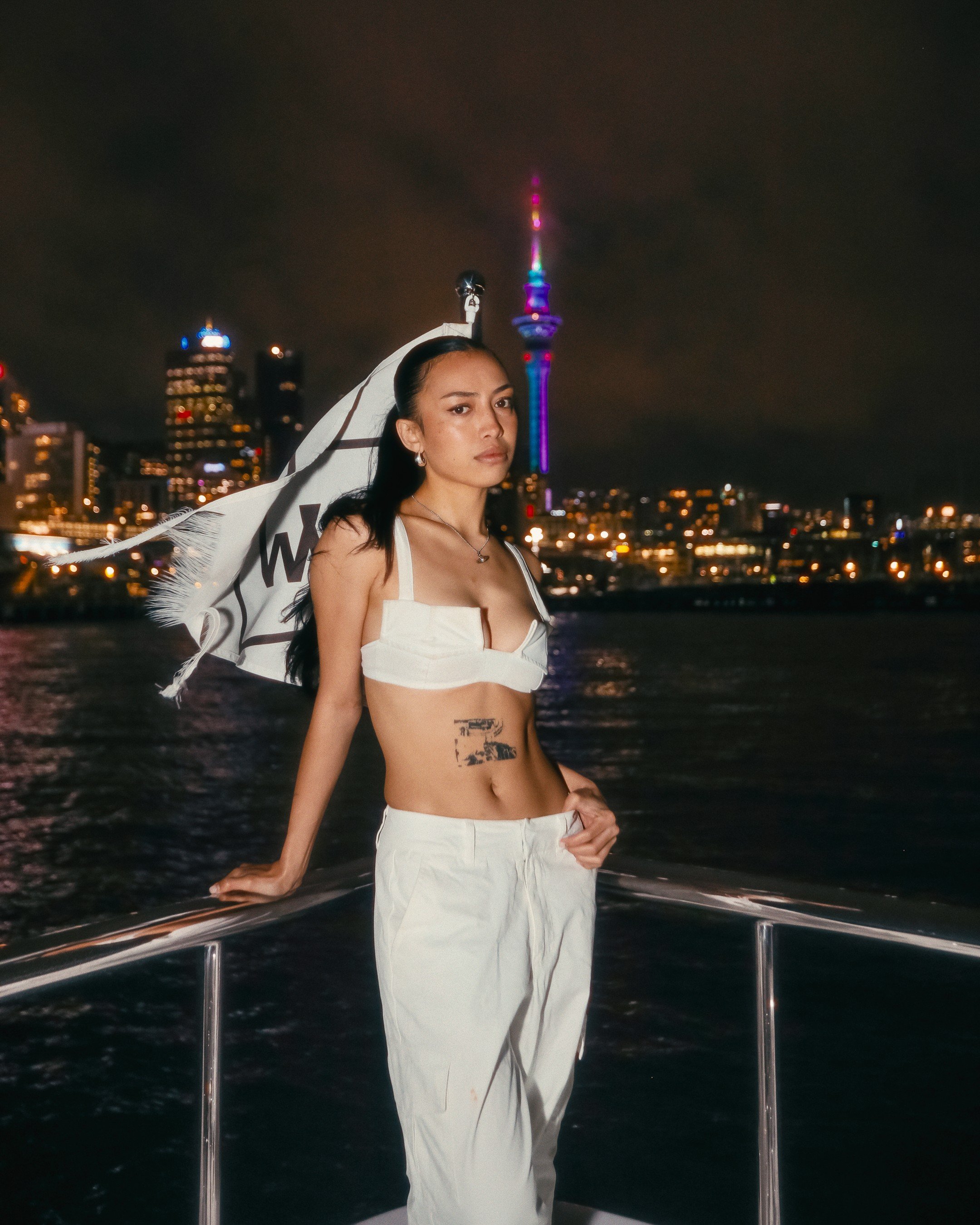 A woman with long dark hair stands on a boat at night, with Auckland city lights and the Sky Tower in the background. She is dressed in white pants and a white cropped top, leaning on the boat's railing.