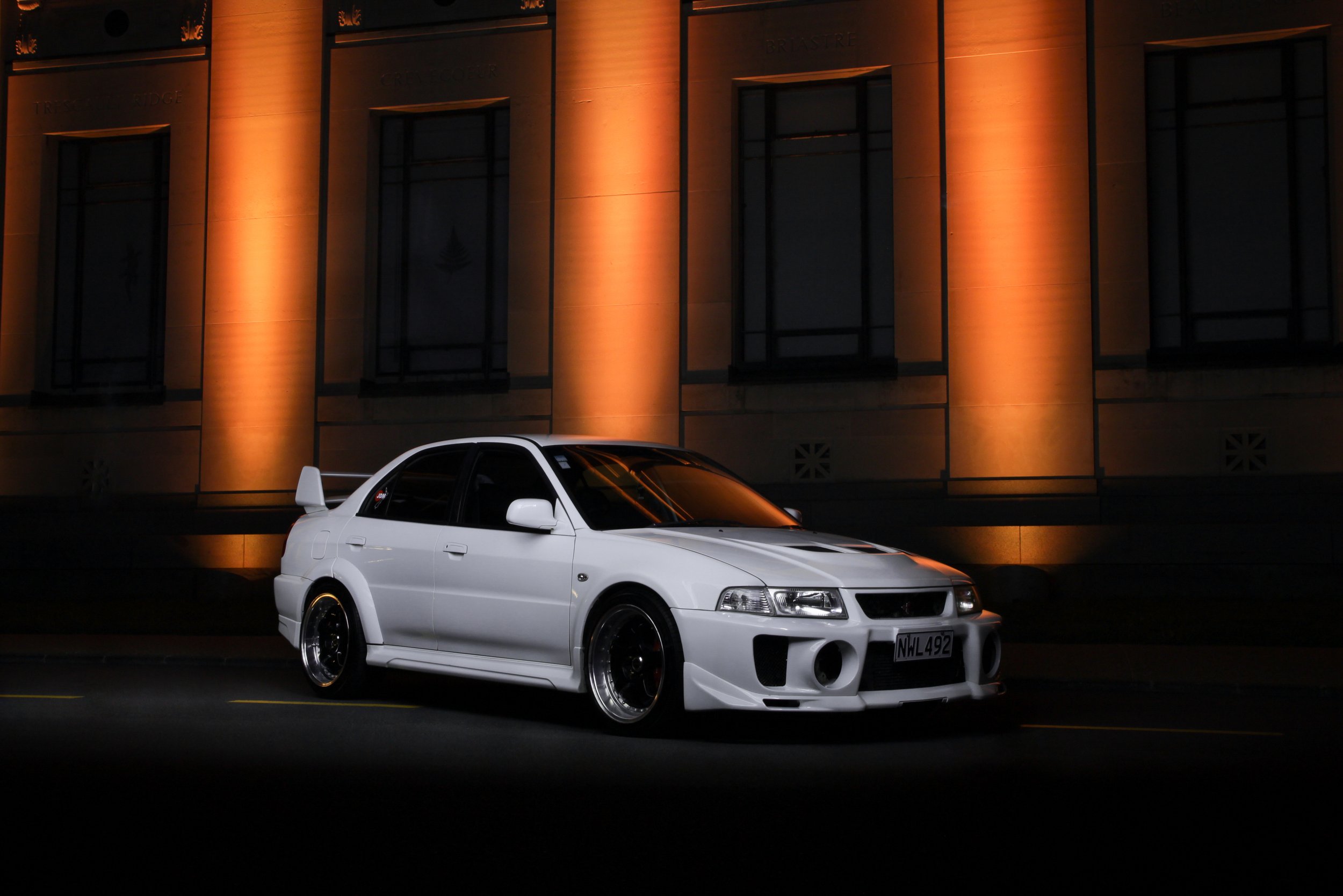 A white Mitsubishi Evo 5 parked in front of an illuminated building at night.