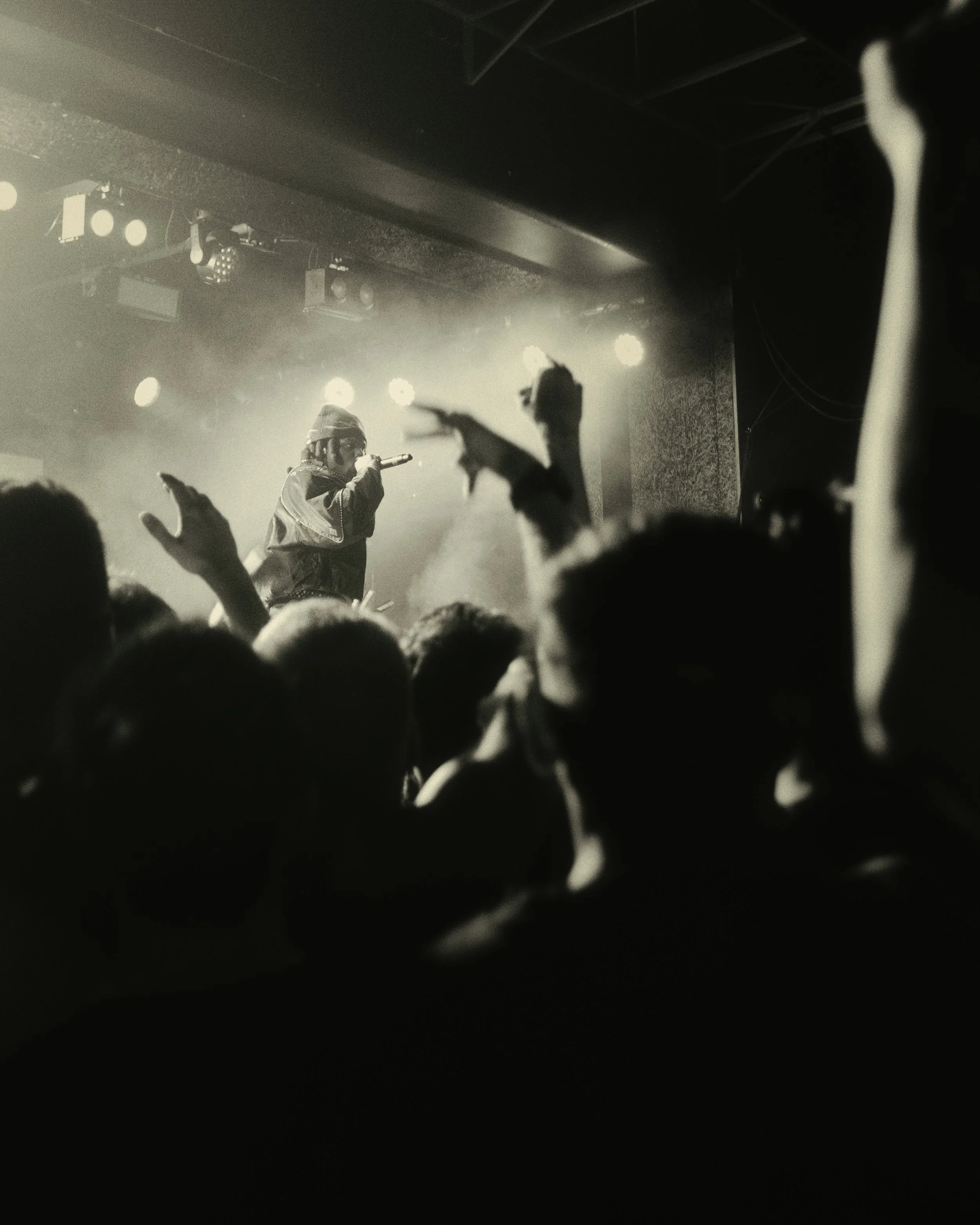 Denzel Curry on stage performing, wearing a jacket and beanie, holding a microphone, with arms raised, and stage lights behind.