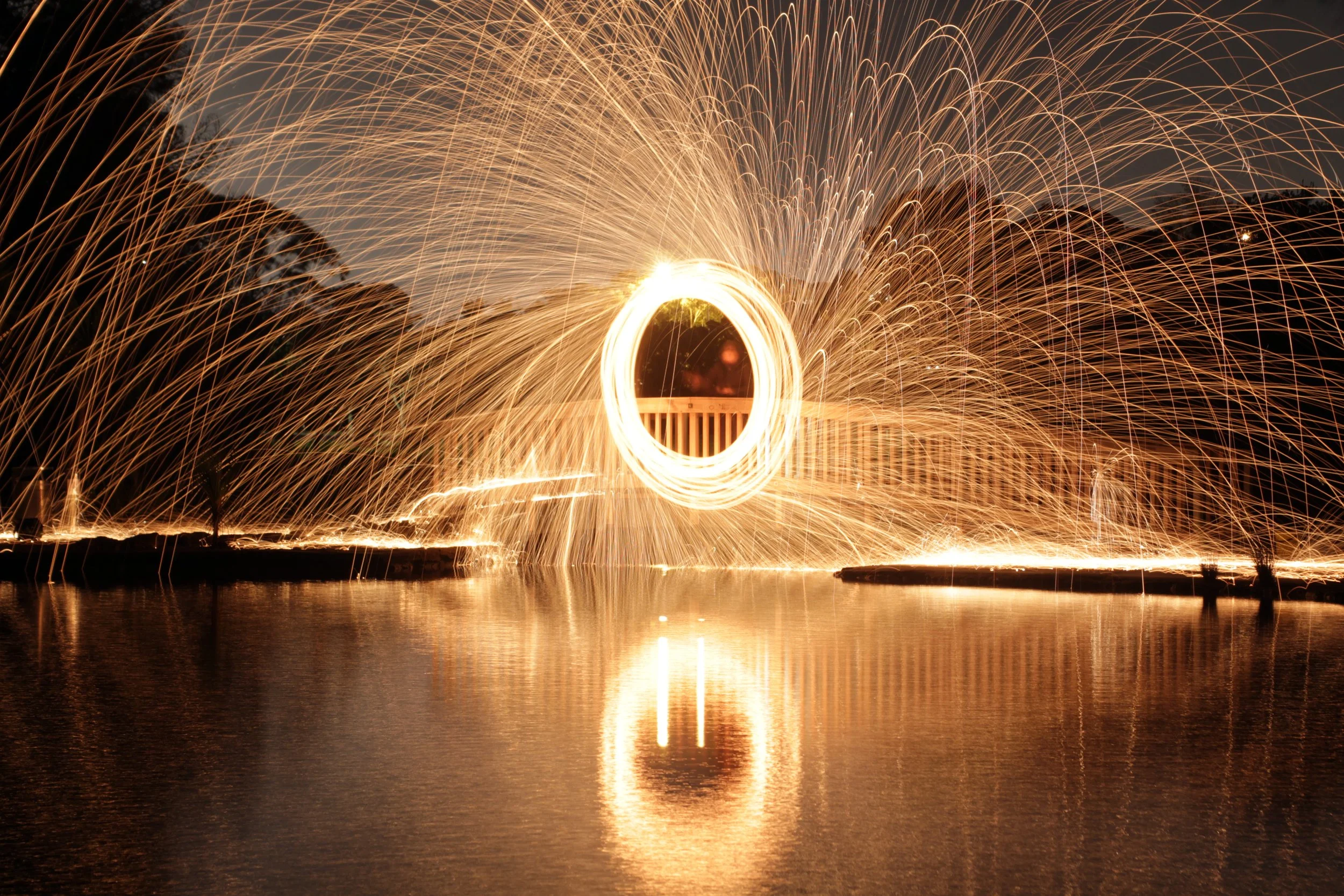 Long exposure photo of steel wool spinning creating sparks, reflected on water, with trees visible in the background at night.