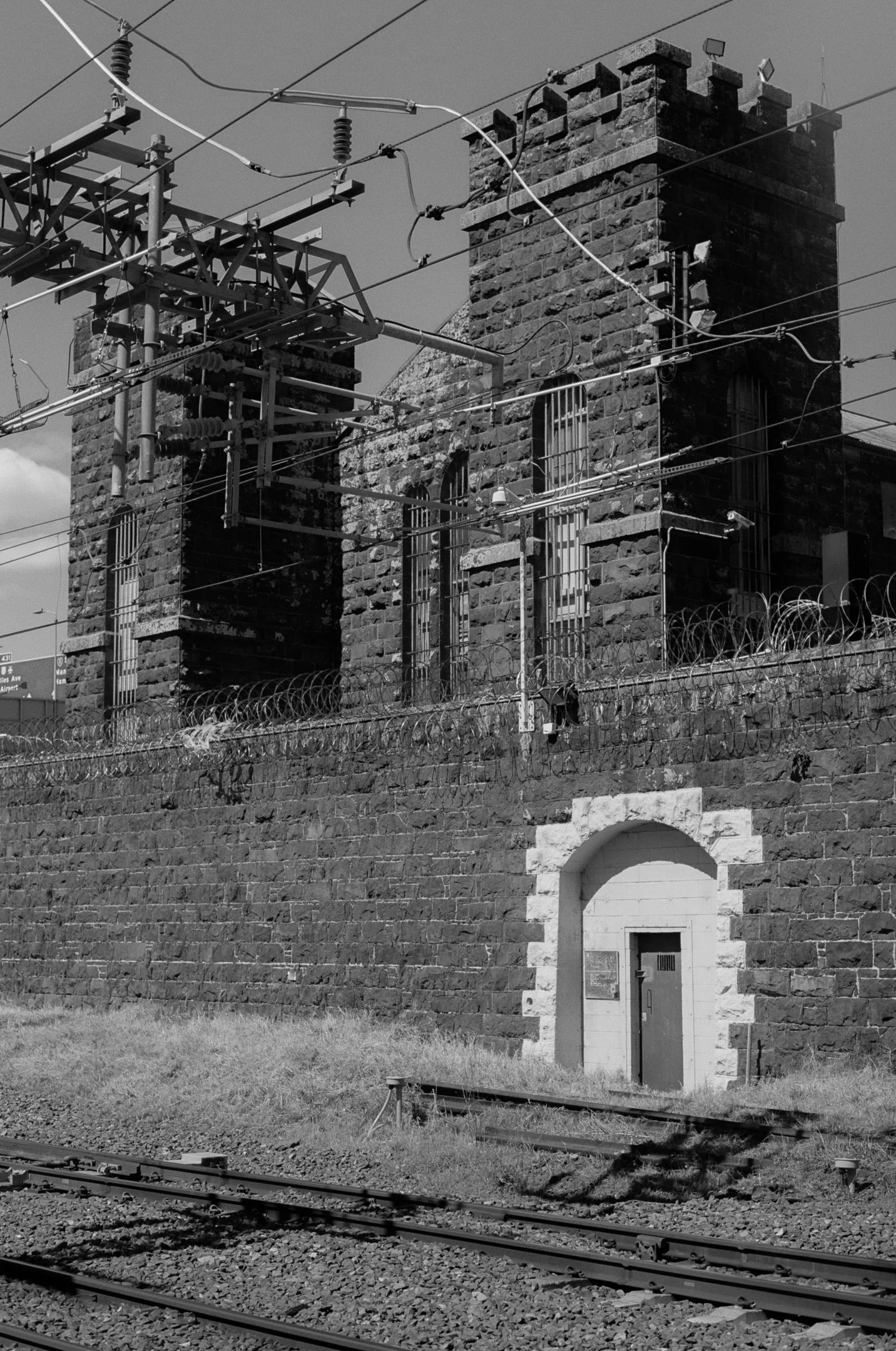 Black and white photo of a brick railway building with a tower, barbed wire fence, and train tracks in the foreground.