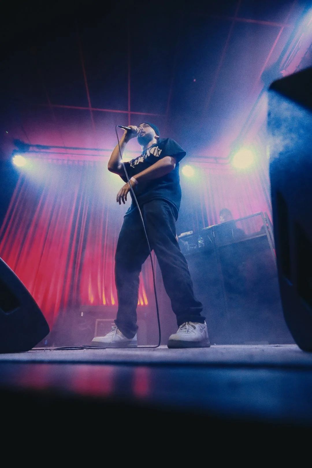 A male performer singing into a microphone on stage with colorful lights and fog, wearing casual clothing and sneakers.