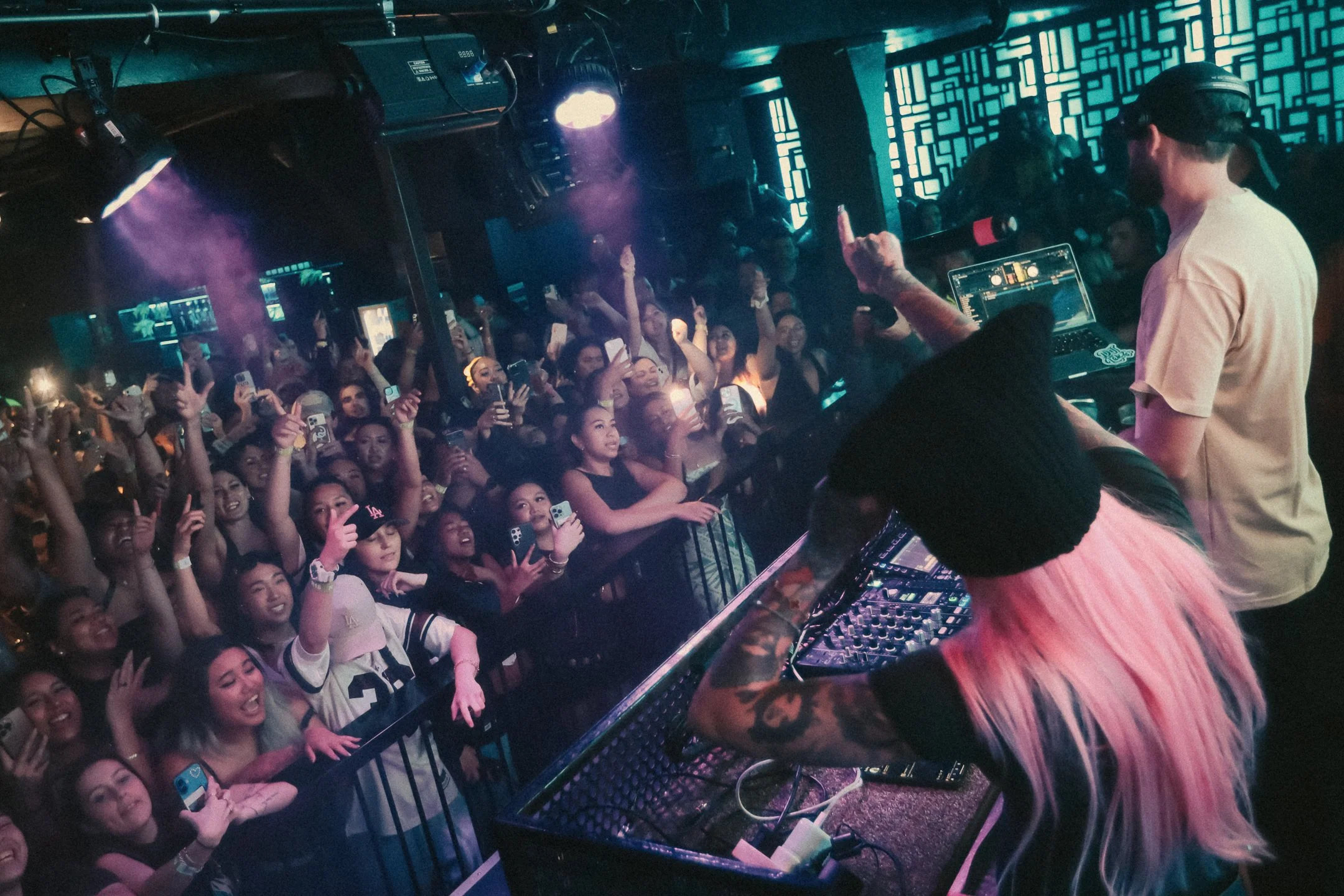Kehlani performs at a nightclub with a crowd dancing and taking photos. Kehlani and the DJ are standing behind a mixing console with colorful lights and smoke effects in the background.