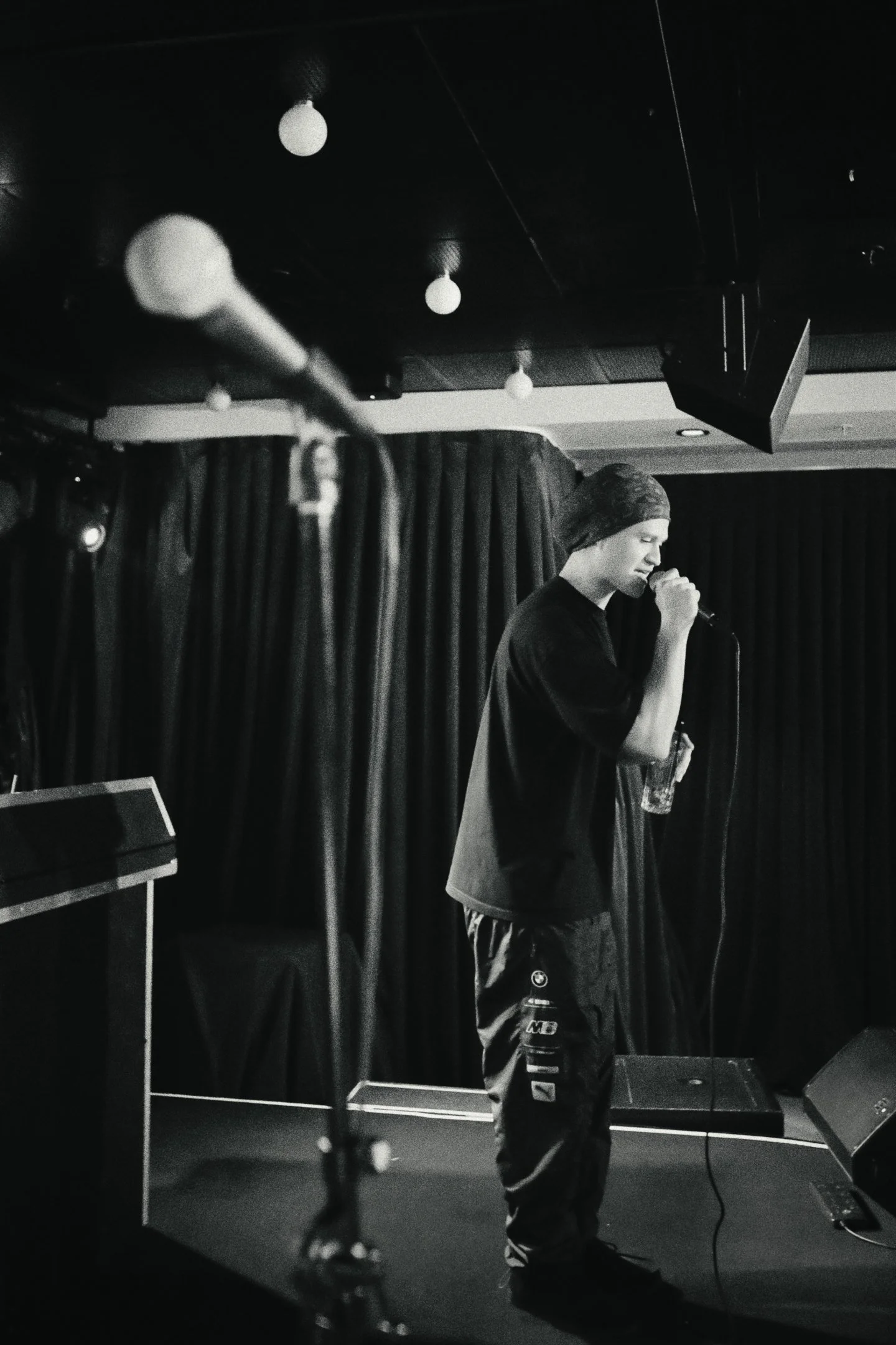 A person, wearing a beanie and sweatpants sings into a microphone, while a microphone stand is in the foreground. There are dark curtains, under ceiling lights and a speaker in the background.