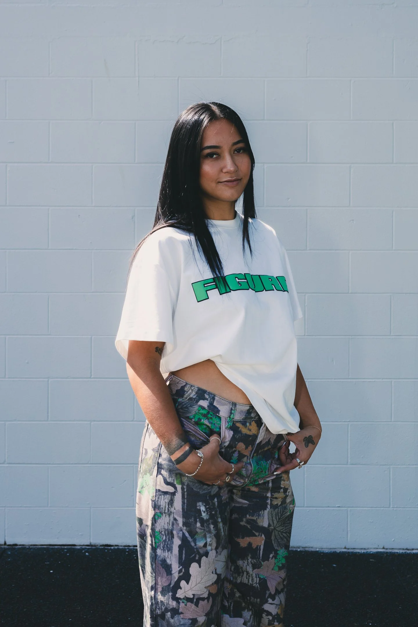 A young woman with long black hair and tattoos standing against a light gray brick wall, wearing a white shirt with 'FIGURE' printed on it and camouflage pants.