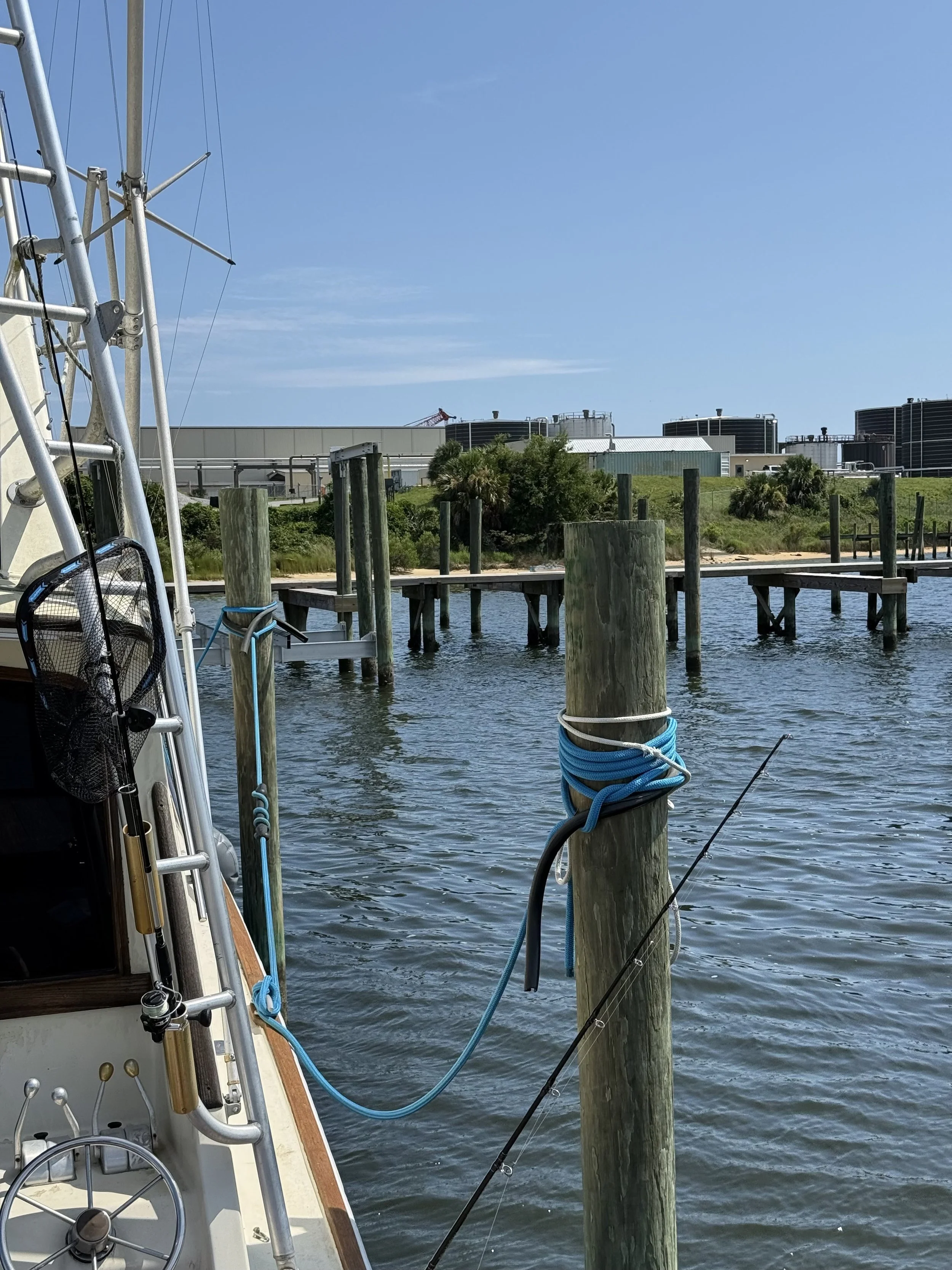 Part of the back of a boat and the piling it is tied to. With water and a second dock in the background.