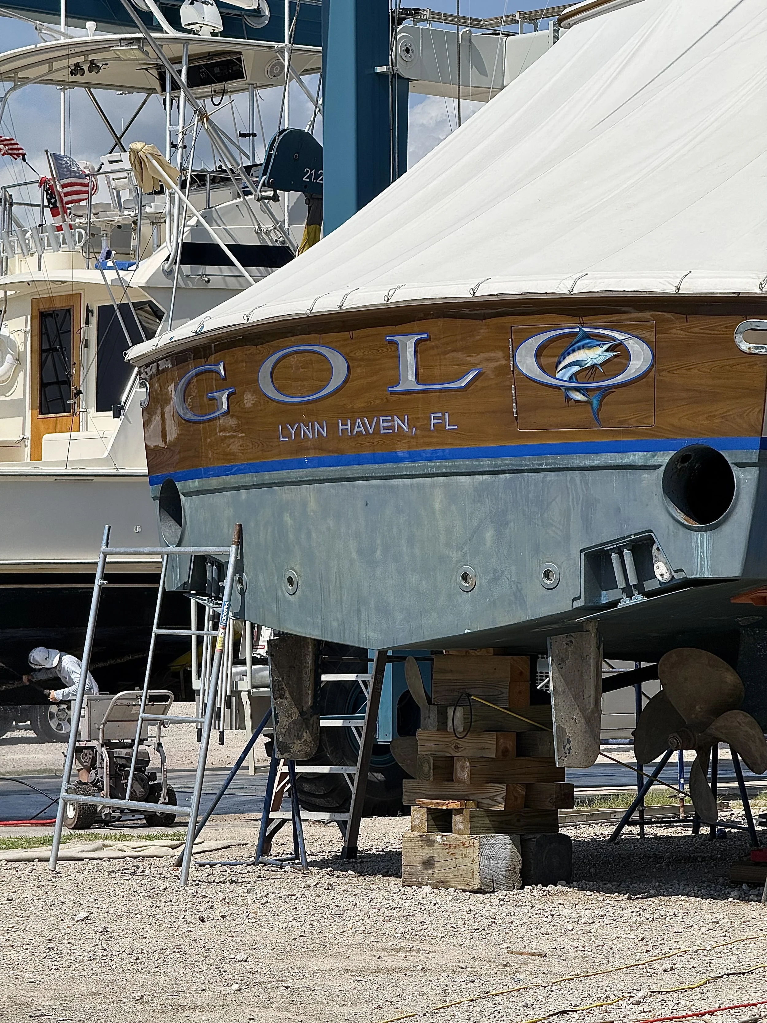A boat named "GOLO" with a blue marlin through the last O lifted up off the ground ready for repairs.