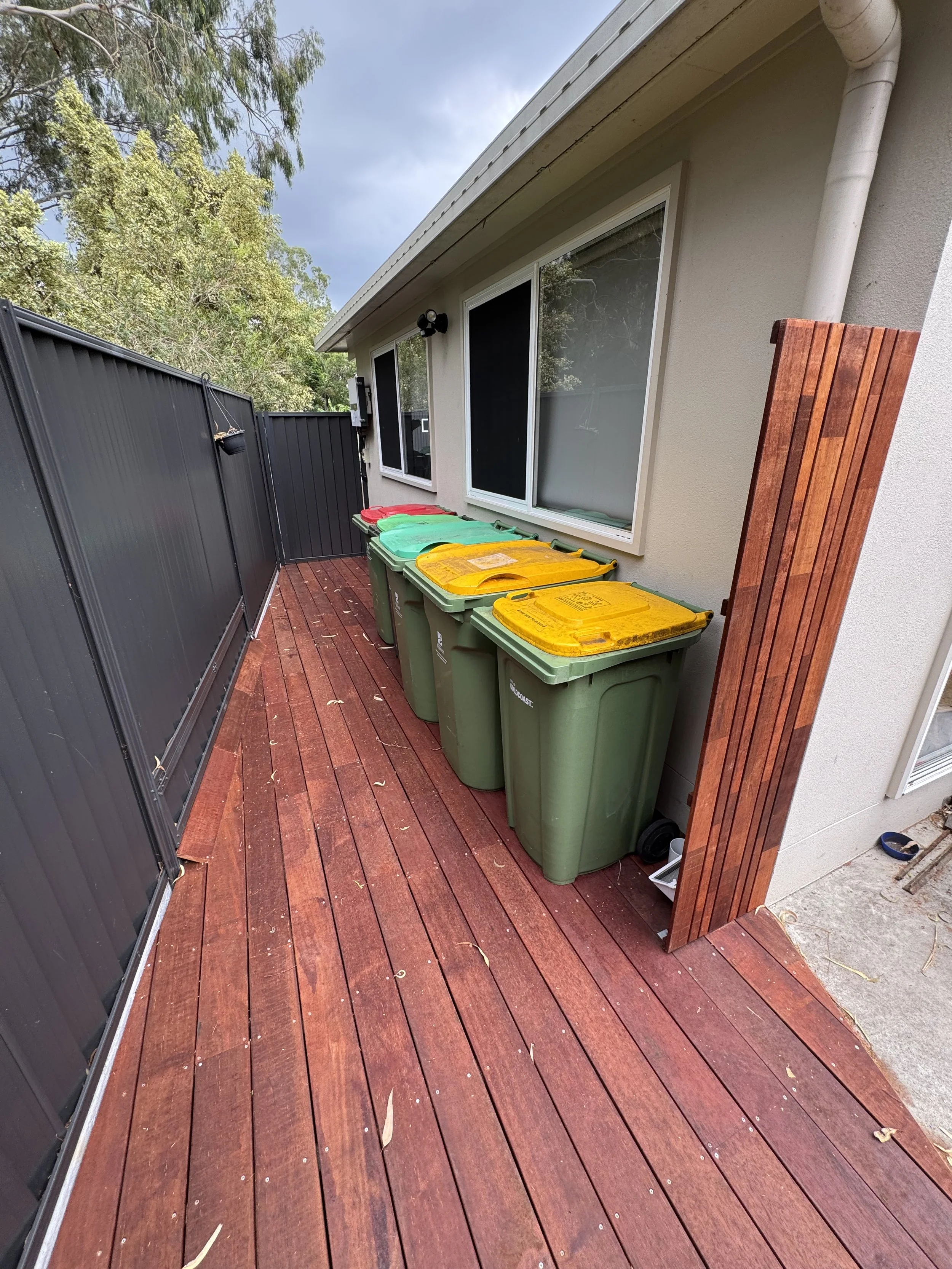 A backyard patio with a wooden deck, black fence, and four green and yellow trash bins lined up against the house wall. There are trees in the background and a cloudy sky.