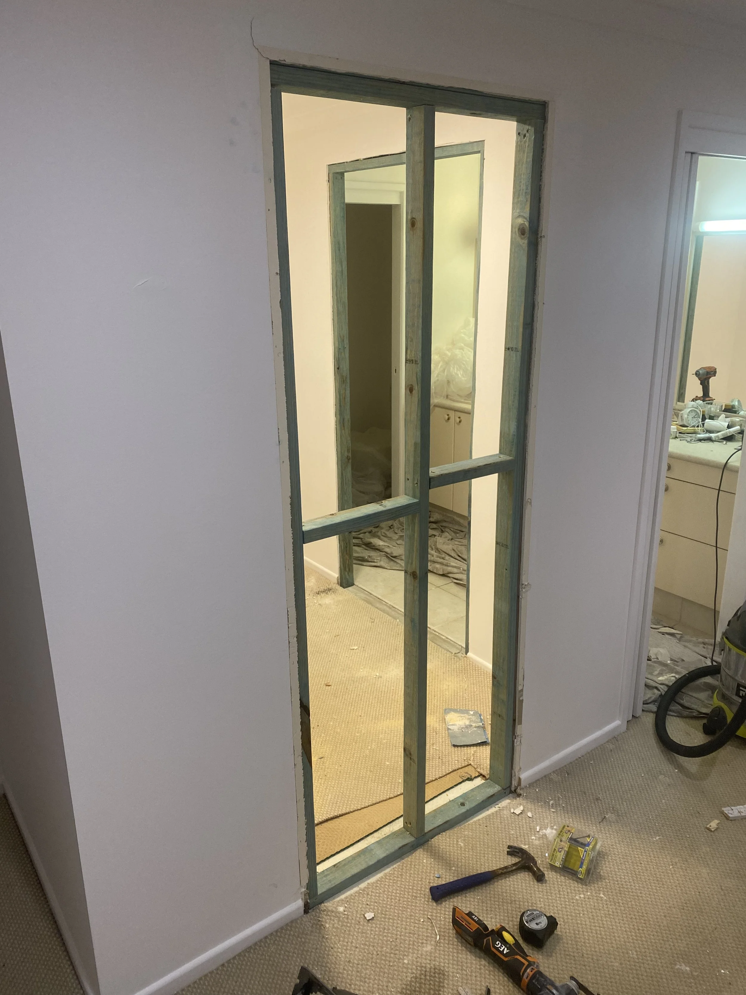 Close-up view of a room under renovation with a partially installed door frame, construction tools including a drill, hammer, measuring tape, and other equipment scattered on the beige carpeted floor. The walls are white and unfinished, and there is visible construction debris.