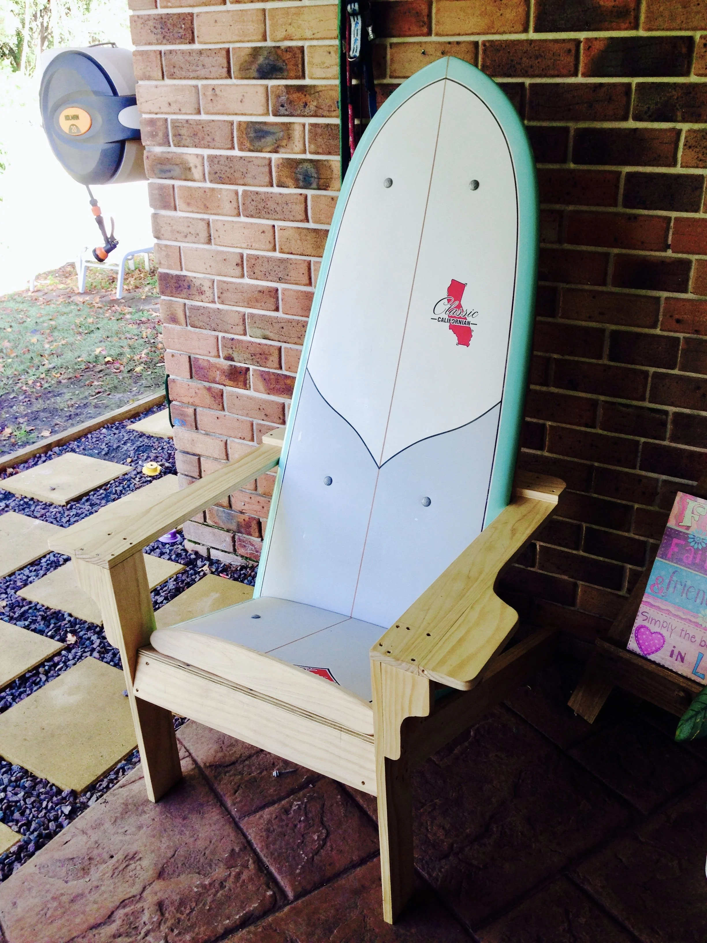 A partially assembled wooden chair featuring a surfboard as the backrest, placed outdoors against a brick wall, with a gravel and paving stone ground nearby.