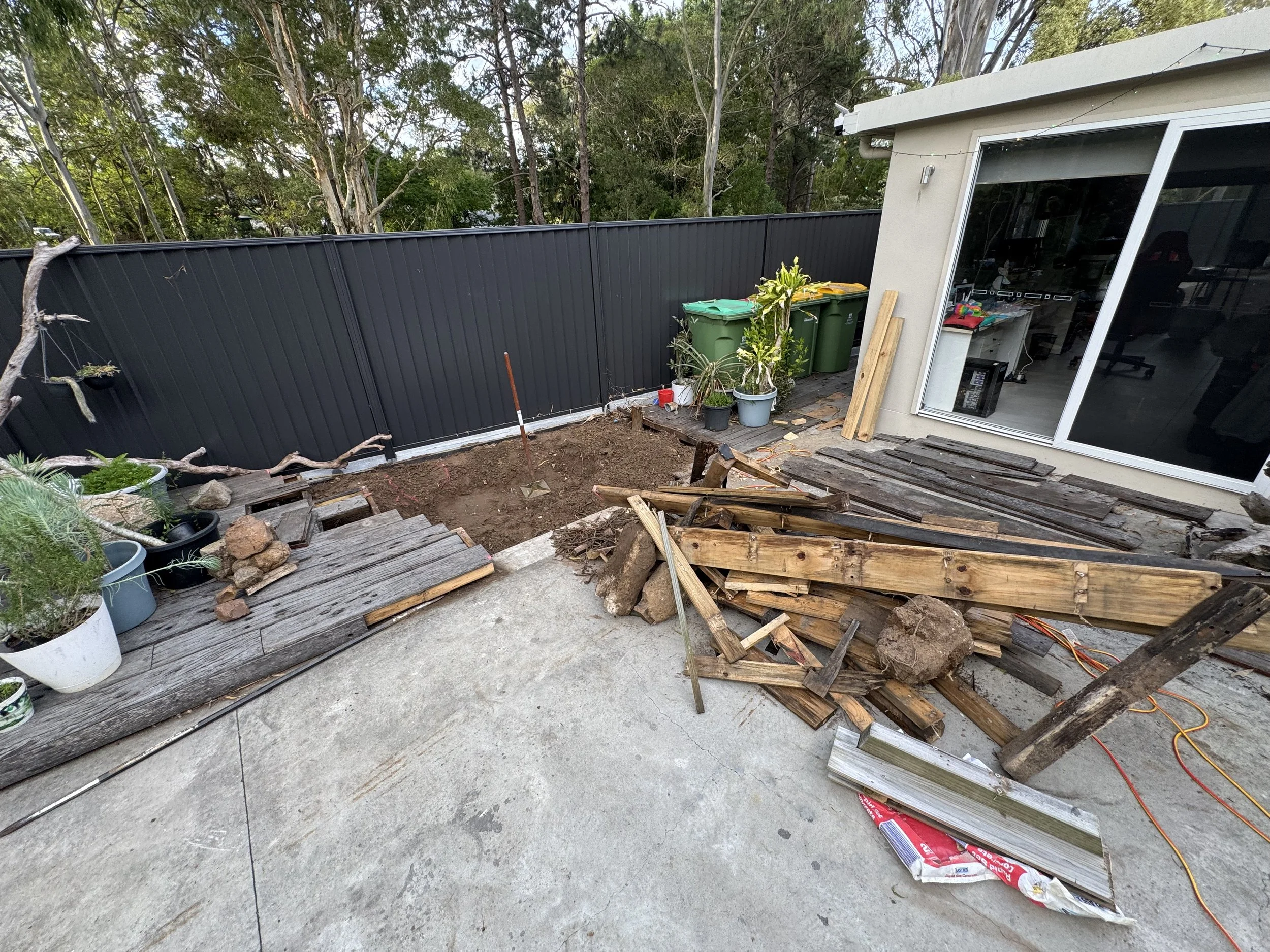 Backyard or patio with construction in progress, including a partially demolished wooden deck, piles of wooden planks, some potted plants, and a black metal fence with green trash bins nearby.