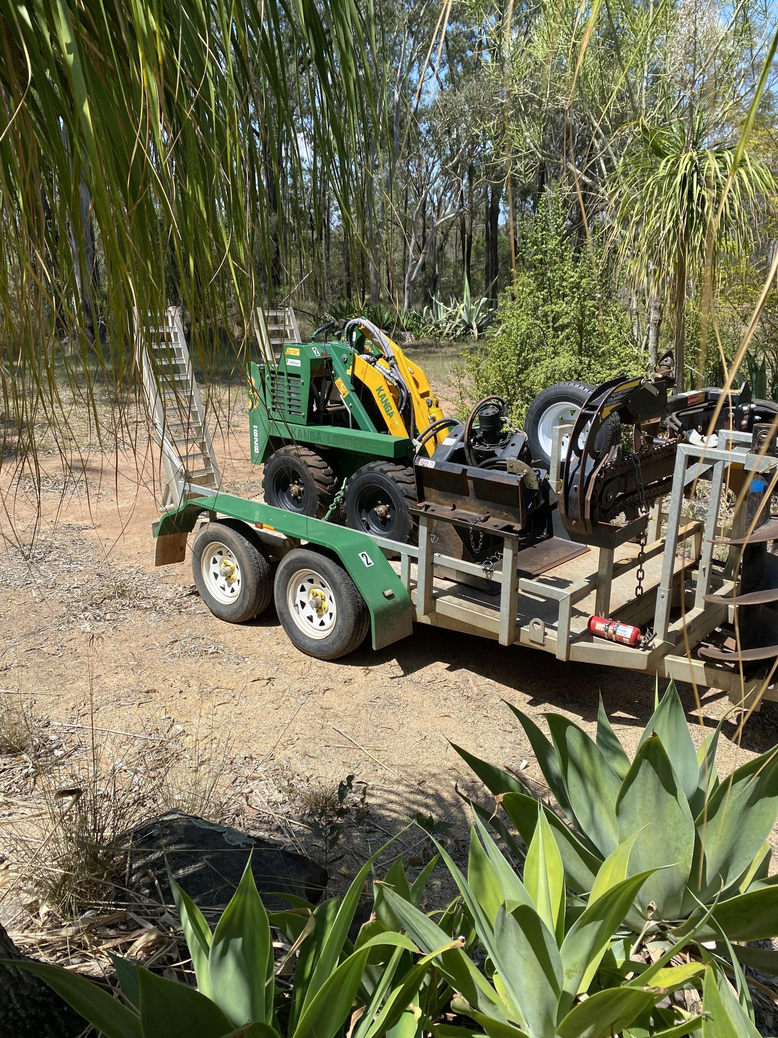 A green and yellow robotic forestry machine on a trailer, parked among trees and plants in a forested area.