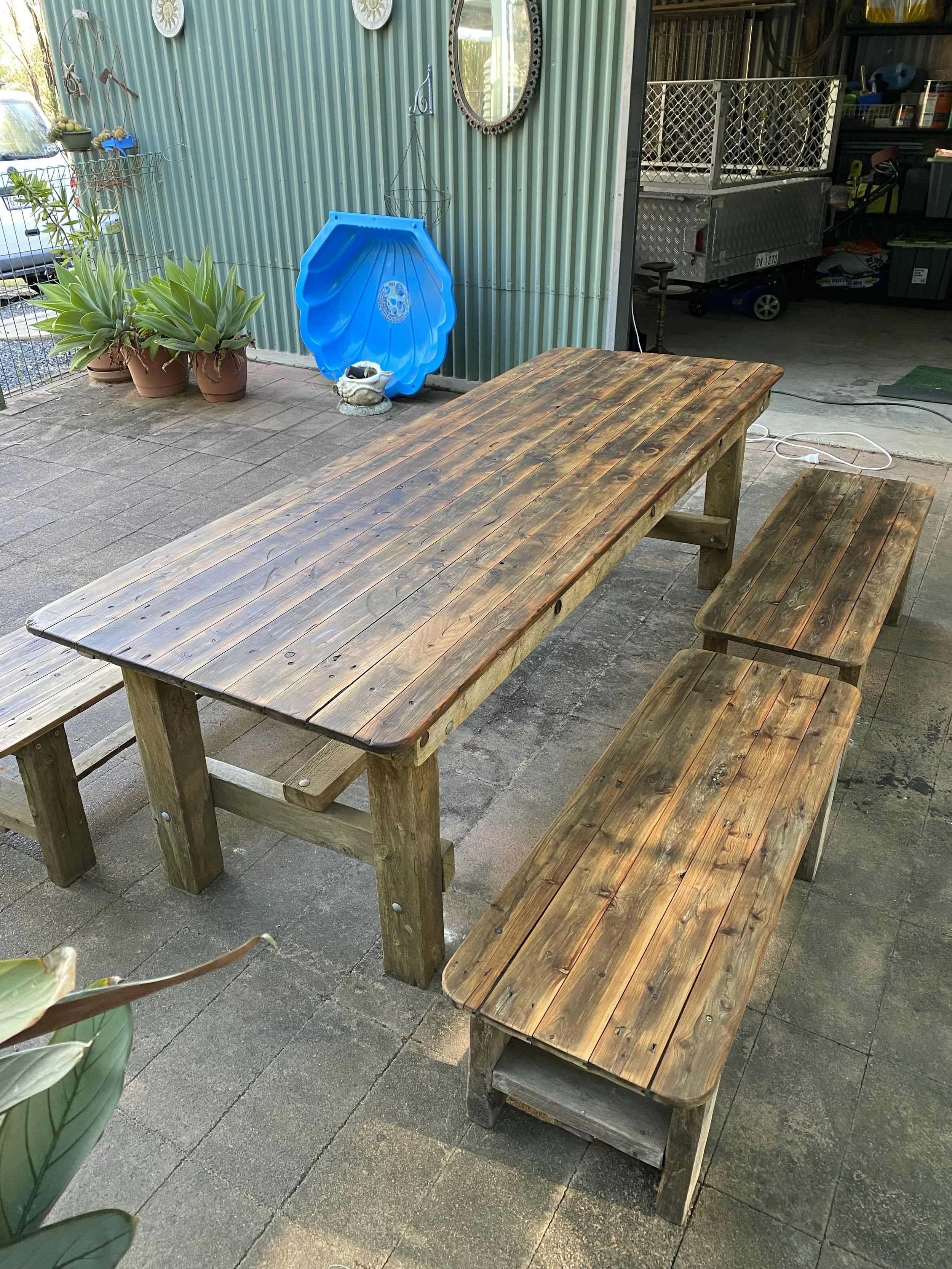 A wooden picnic table and benches on a stone patio, with potted plants and gardening tools visible in the background.