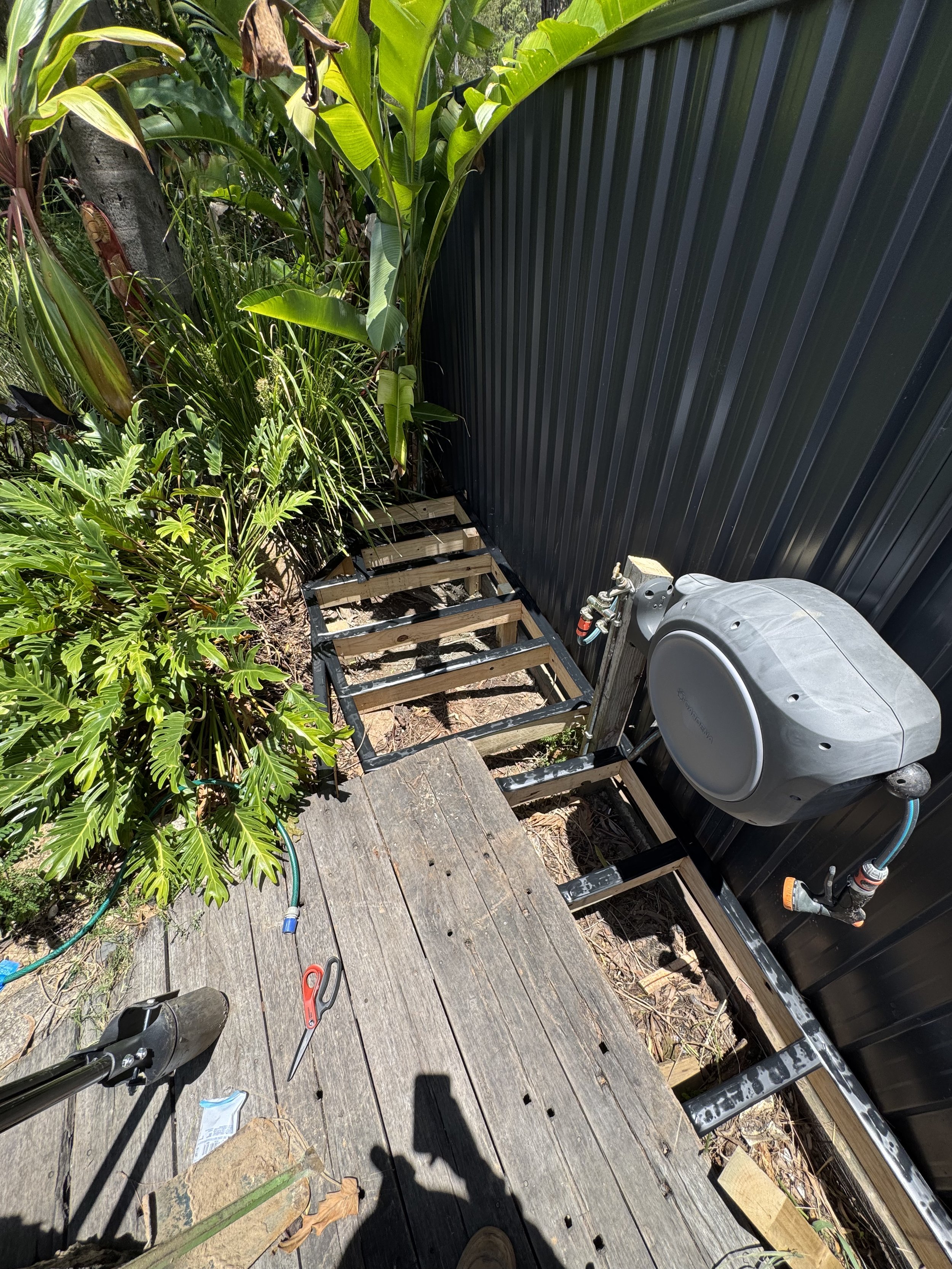 Outdoor deck with tools and gardening supplies, including scissors, a garden hose, and a fan, surrounded by green plants and a black metal fence.