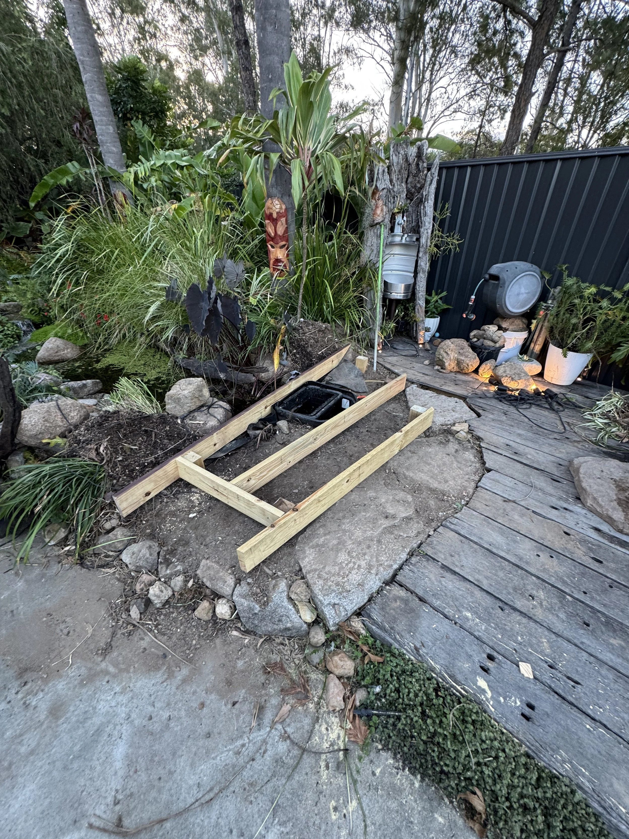 A garden scene with plants, a wooden frame on the ground, rocks, a black fence, and garden tools and decorations, including a water barrel and masks on a fence post.