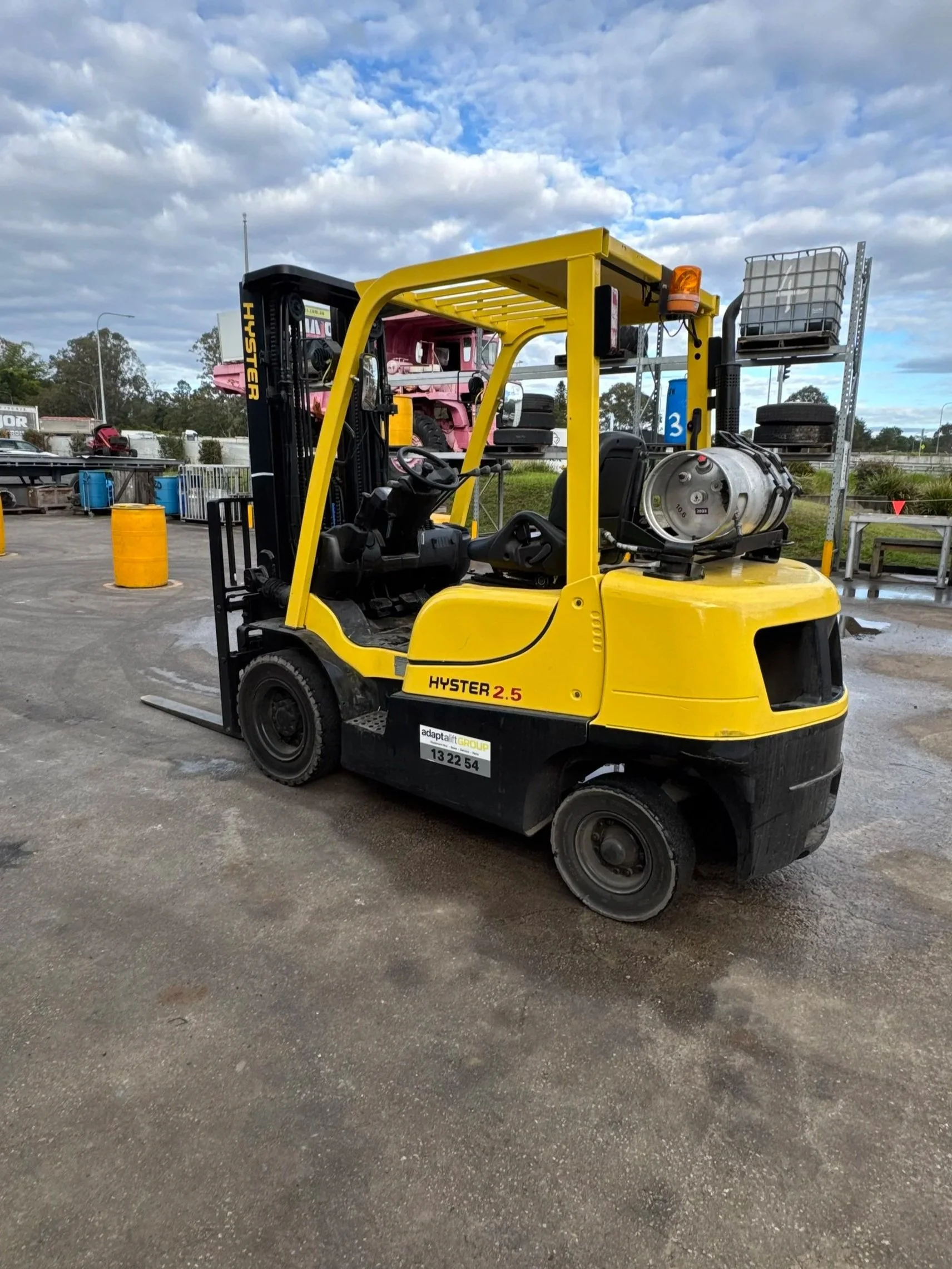 Yellow forklift with a propane tank onboard, parked outdoors under a partly cloudy sky with industrial and natural surroundings.