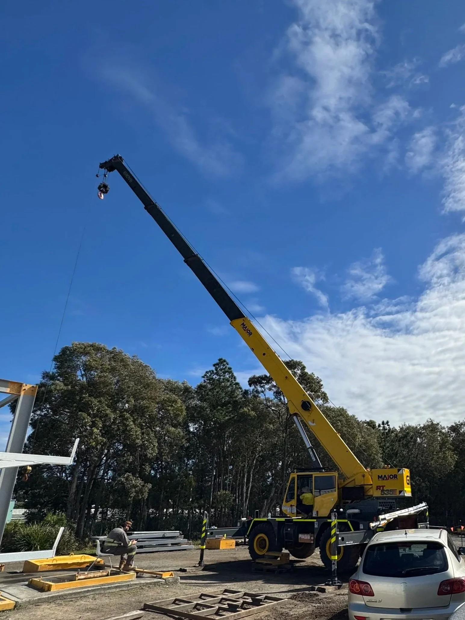 Construction crane lifting materials at a construction site with workers and vehicles, trees in the background, under a partly cloudy sky.