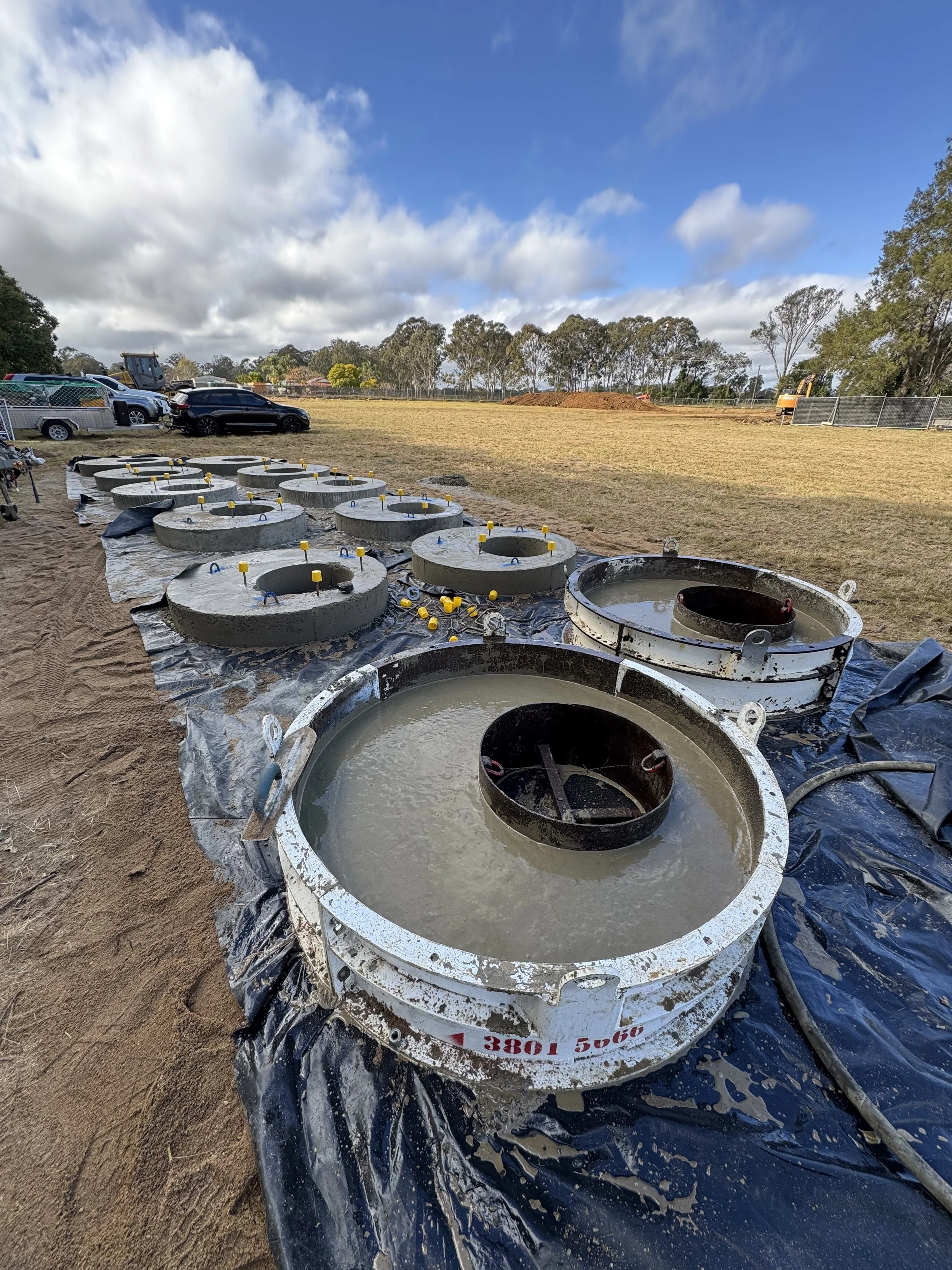 Construction site with concrete cylinders filled with liquid, metal rings, parked cars and construction vehicles on a sandy ground under a partly cloudy sky.