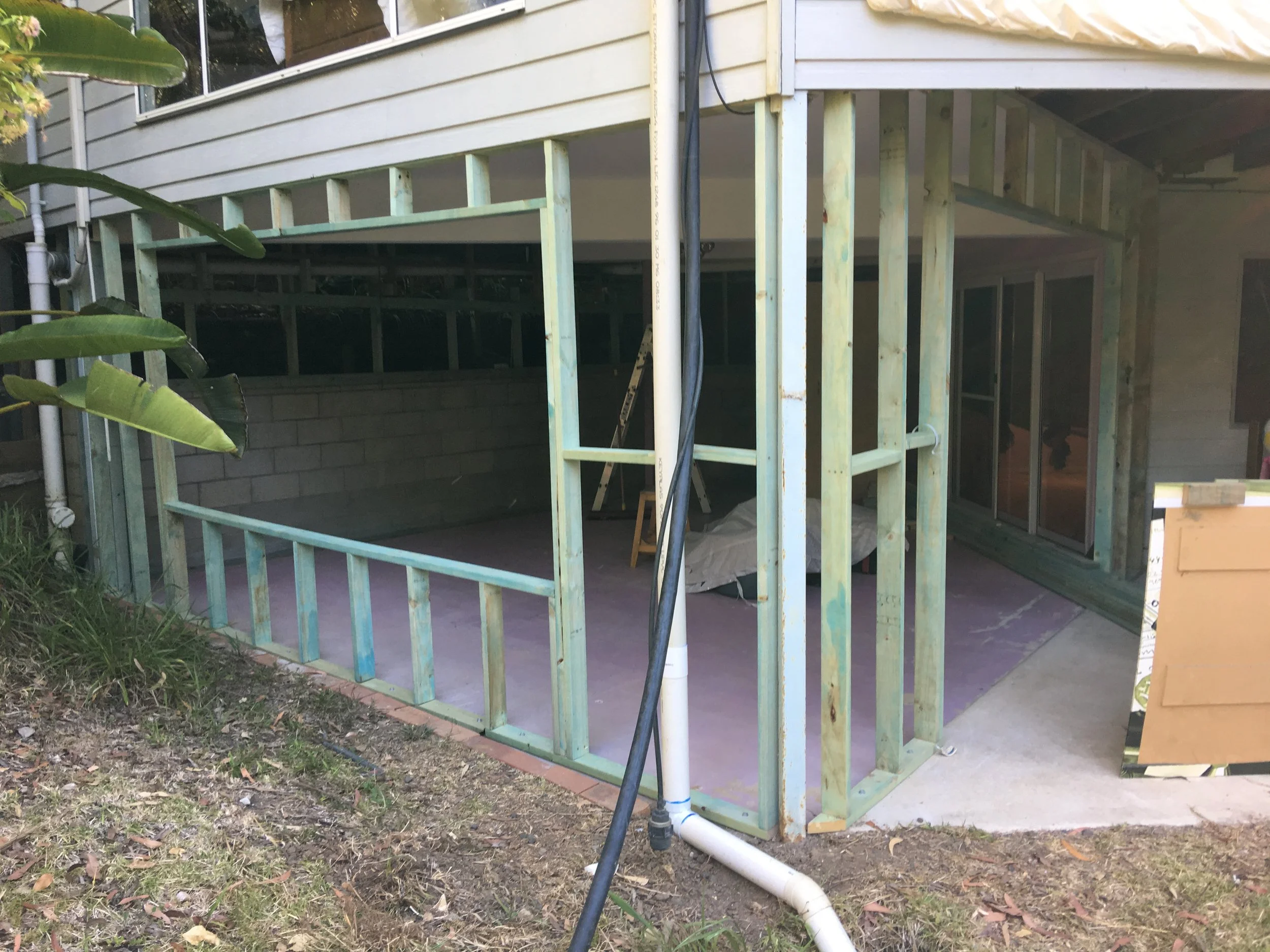 Exterior view of a house under construction, showing the wooden framing of a deck or porch with some parts painted light green. Construction materials and tools are visible, along with black and white PVC pipes and black electrical wiring, with green plants nearby.