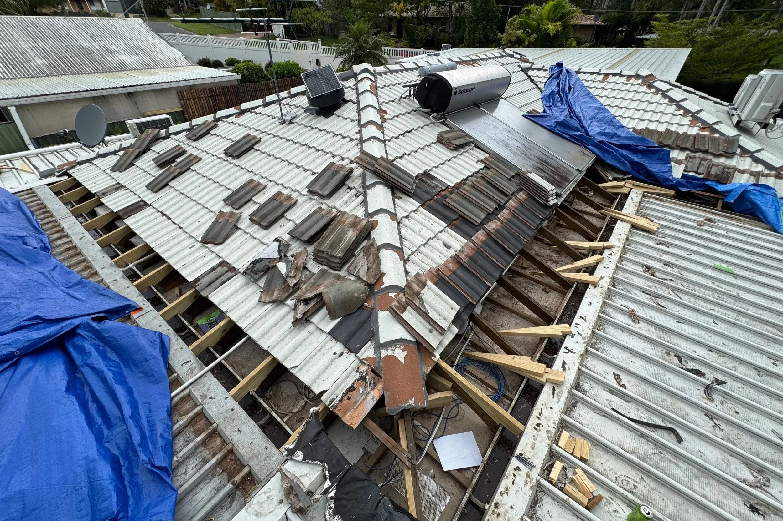The image shows a rooftop undergoing repair or renovation, with some tiles removed and work in progress. Construction materials and tools are visible, including a blue tarp, wooden supports, and solar water heater equipment. The surrounding area has trees and neighboring houses.