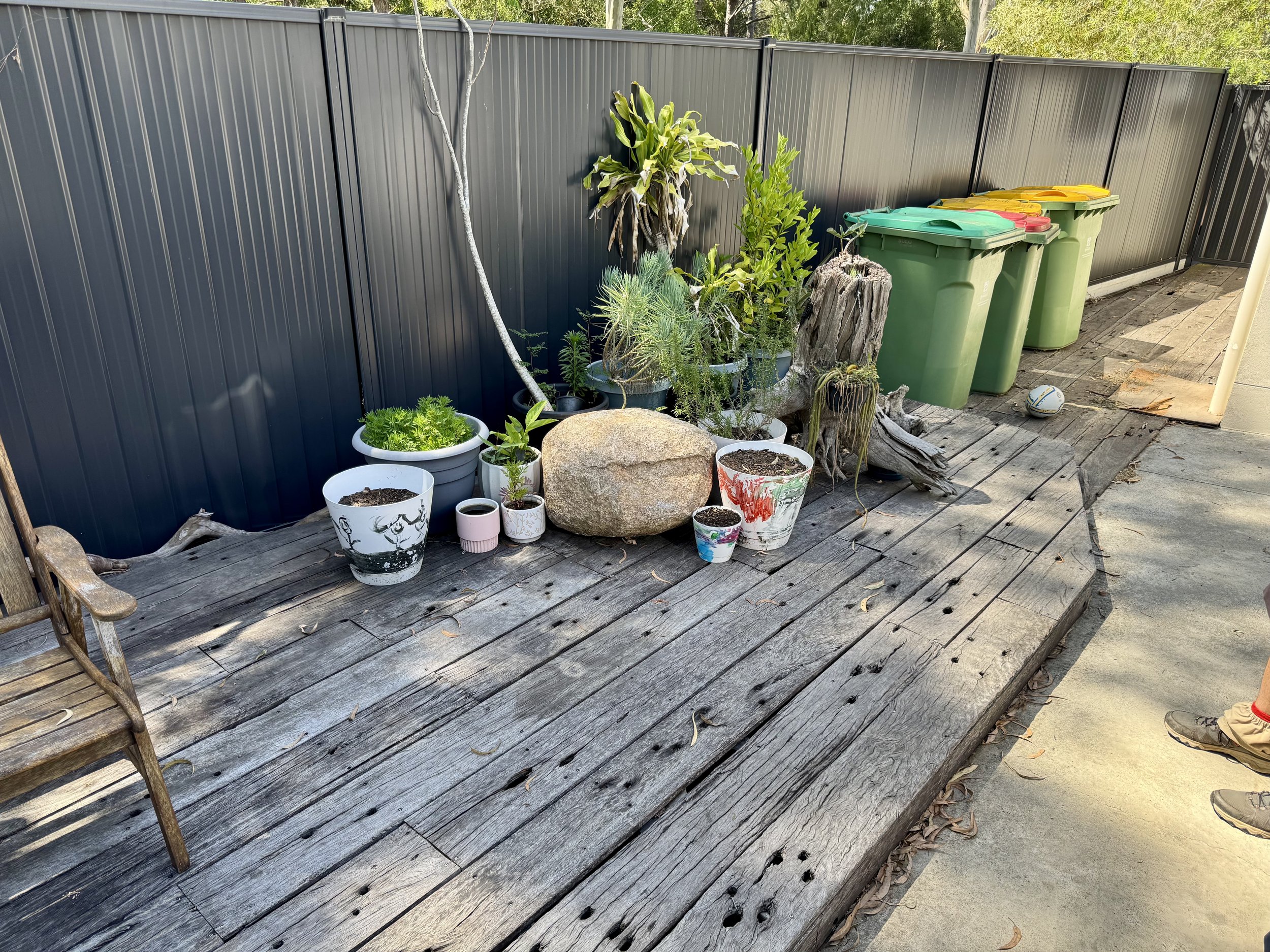 A small outdoor patio area with a wooden deck, plants in pots, a large rock, a weathered tree stump, and a black metal fence. Three green trash cans with yellow, green, and red lids are lined up against the fence, and a ball is on the ground near a person's shoe.