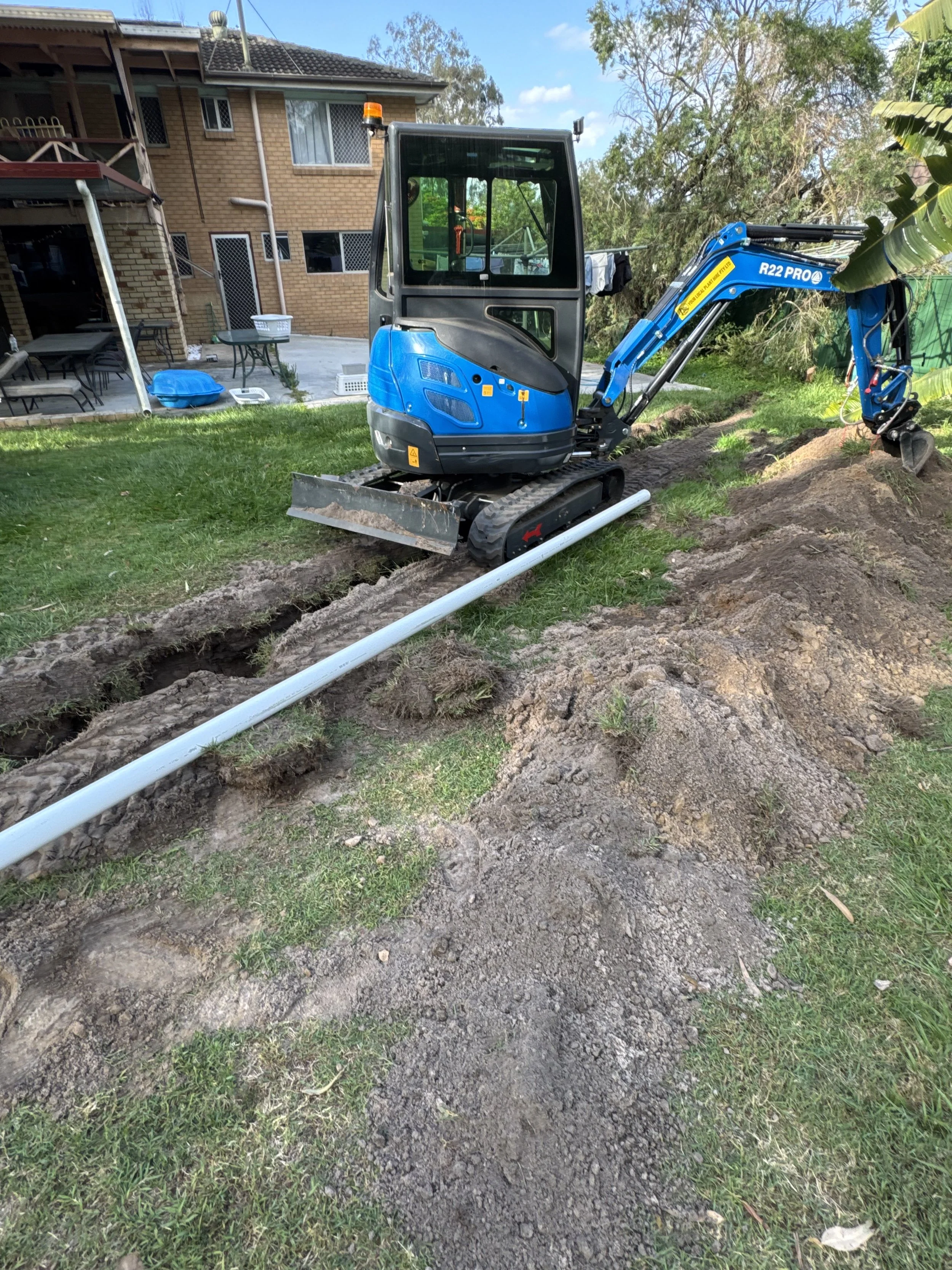A blue and black mini excavator digging a trench in a backyard with a green lawn, near a multi-story brick house and trees.