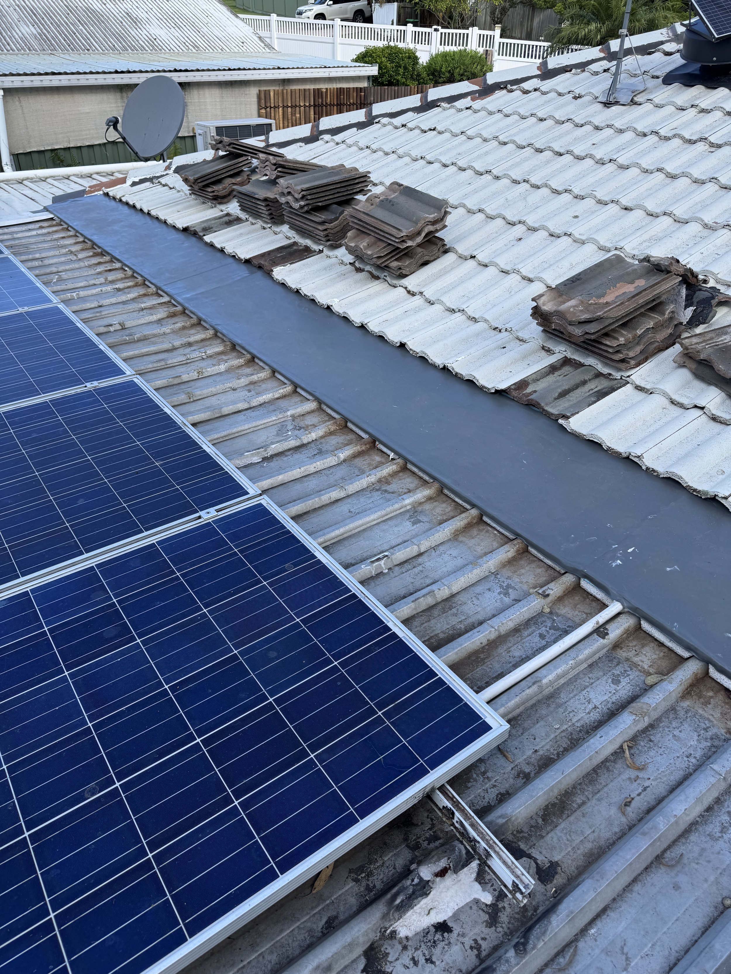 Roof with solar panels installed, stacks of old roof tiles, and a covered vent pipe, with neighboring roofs and a fenced yard in the background.