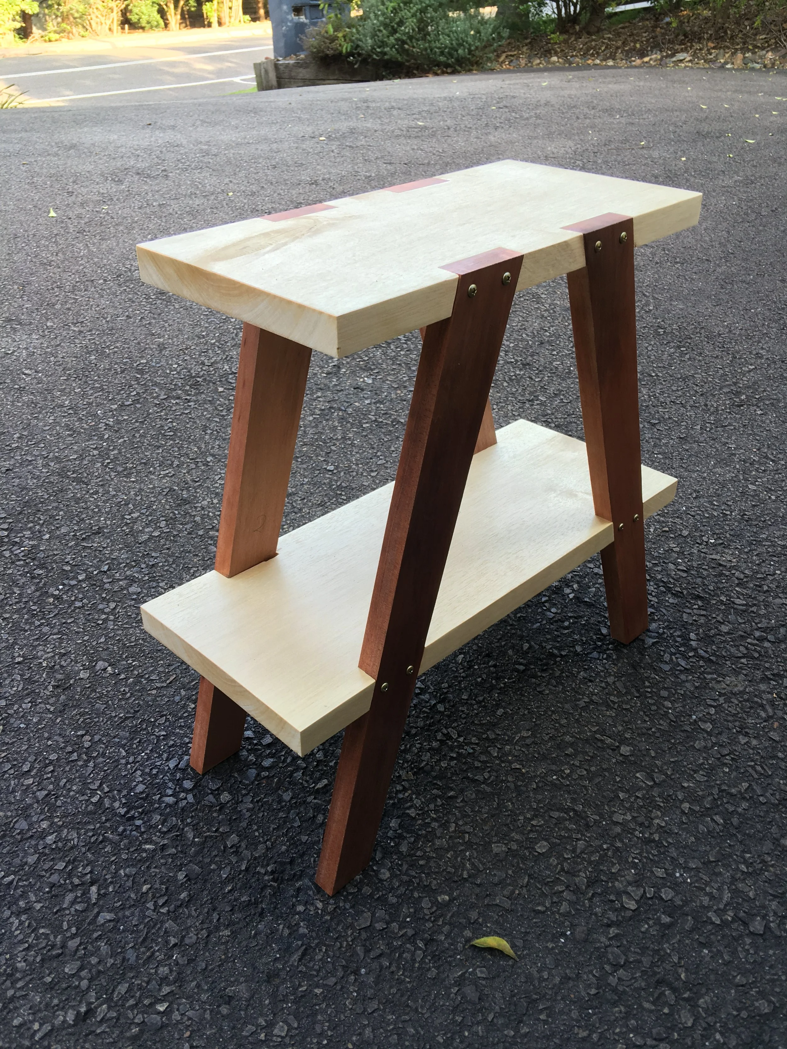 A wooden tiered plant stand with a light-colored top shelf and a darker bottom shelf, supported by diagonally placed wooden legs, situated outdoors on a paved surface with some foliage visible in the background.